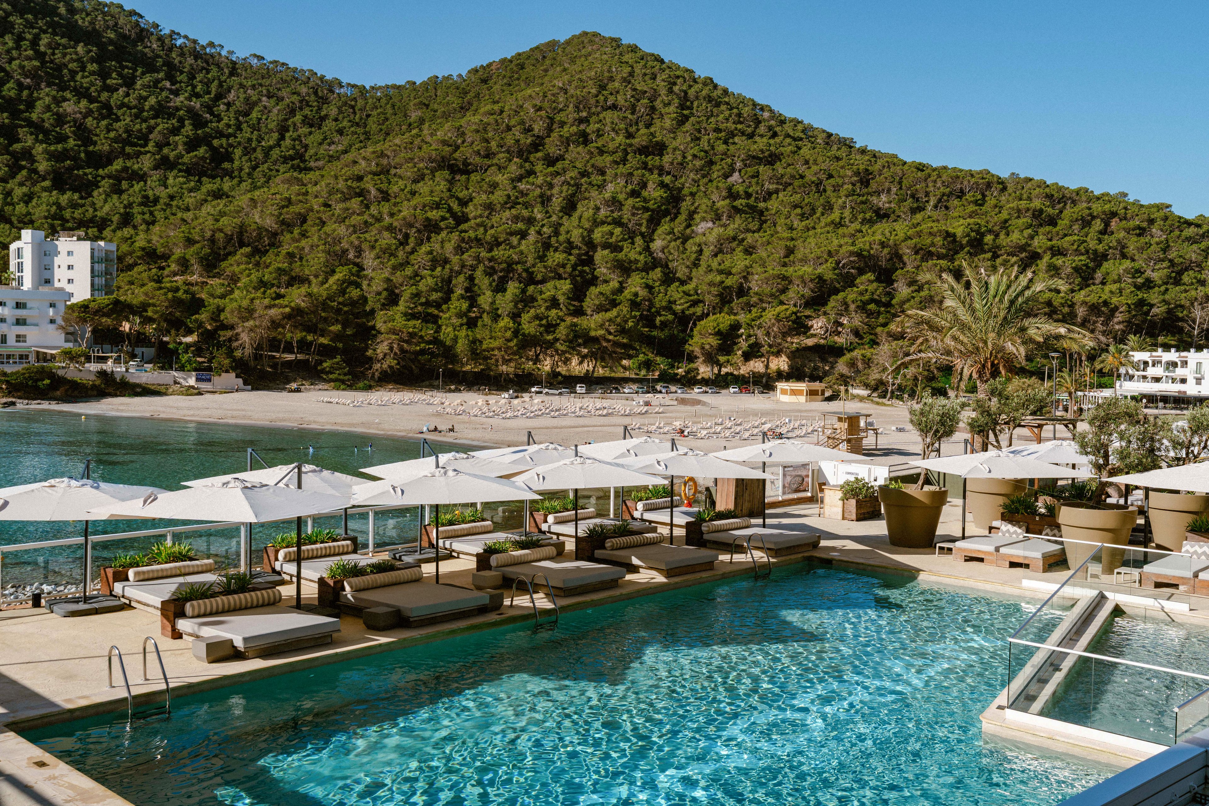 A turquoise outdoor swimming pool overlooks a sandy beach and calm bay. Sun loungers and umbrellas line the poolside terrace, creating a relaxed vacation atmosphere. Lush green hills rise in the background, with a few white buildings near the shoreline. The scene suggests a sunny coastal resort ideal for leisure and relaxation.