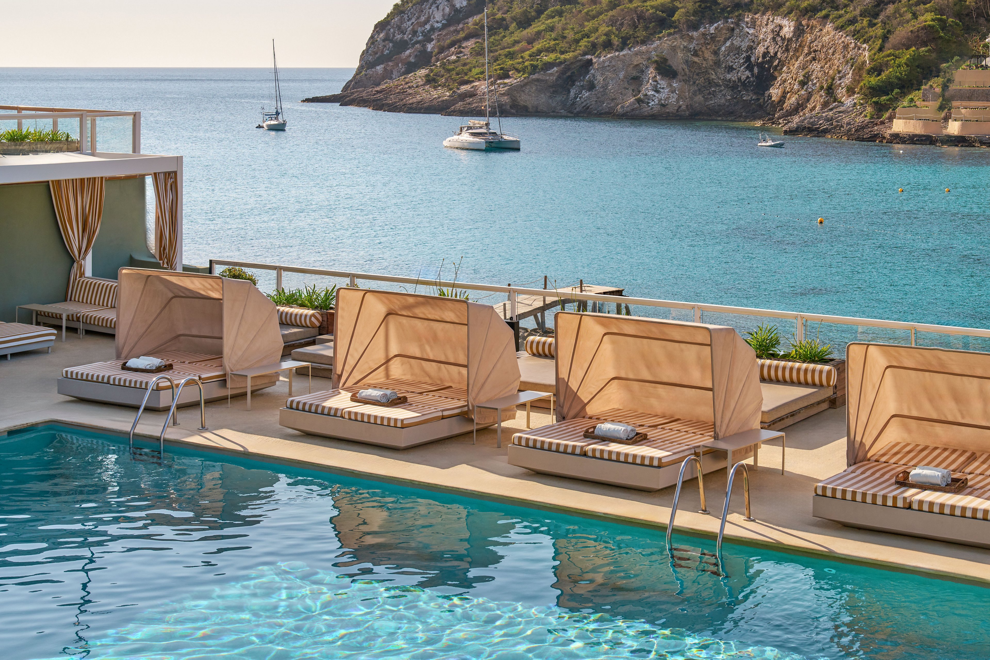 A stylish outdoor pool terrace overlooks a calm turquoise sea and rocky coastline. Beige cabanas and cushioned lounge chairs line the poolside, creating an upscale resort atmosphere. The water in the pool sparkles in the sunlight, while sailboats float in the bay beyond. Neutral tones and clean lines give the scene a modern, relaxing coastal vibe.
