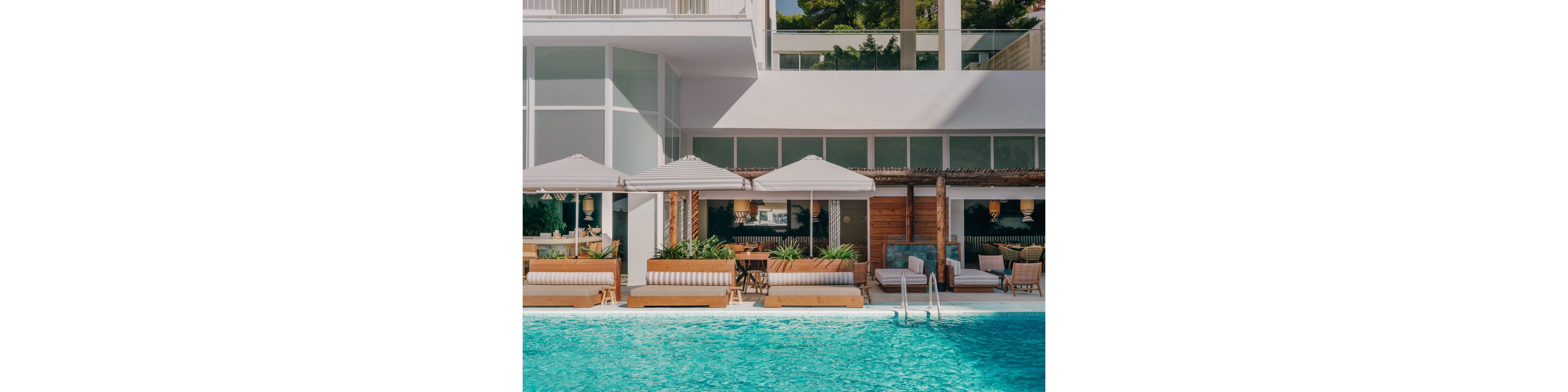 A turquoise swimming pool sits in front of a stylish resort lounge area. Cushioned sunbeds and cabanas line the poolside, accented by potted greenery and wooden paneling. Large windows and white architectural elements create a clean, contemporary look. The scene feels calm and inviting, suggesting a relaxed vacation atmosphere in warm weather.