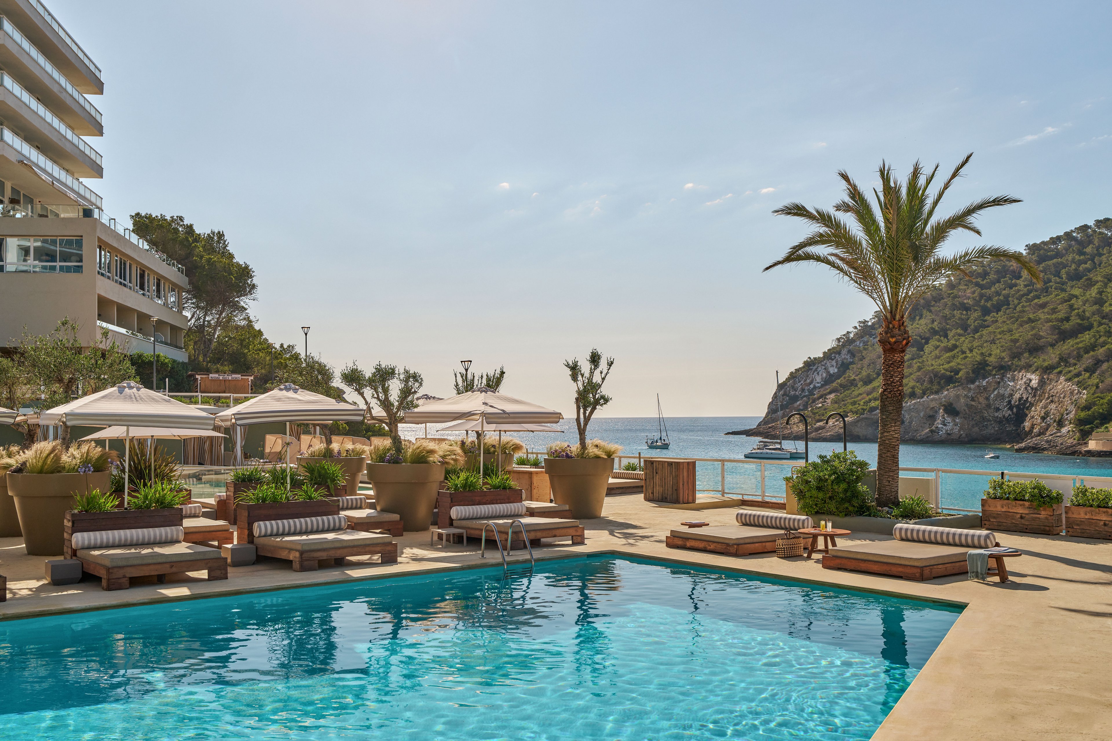 An outdoor swimming pool sits in front of a modern seaside hotel overlooking a calm bay. Sun loungers and cushioned daybeds are arranged around the pool, shaded by large white umbrellas and surrounded by potted plants. A tall palm tree and green hillside frame the view toward the ocean, where a sailboat is anchored in the distance. The scene suggests a tranquil, upscale vacation setting under clear, sunny skies.