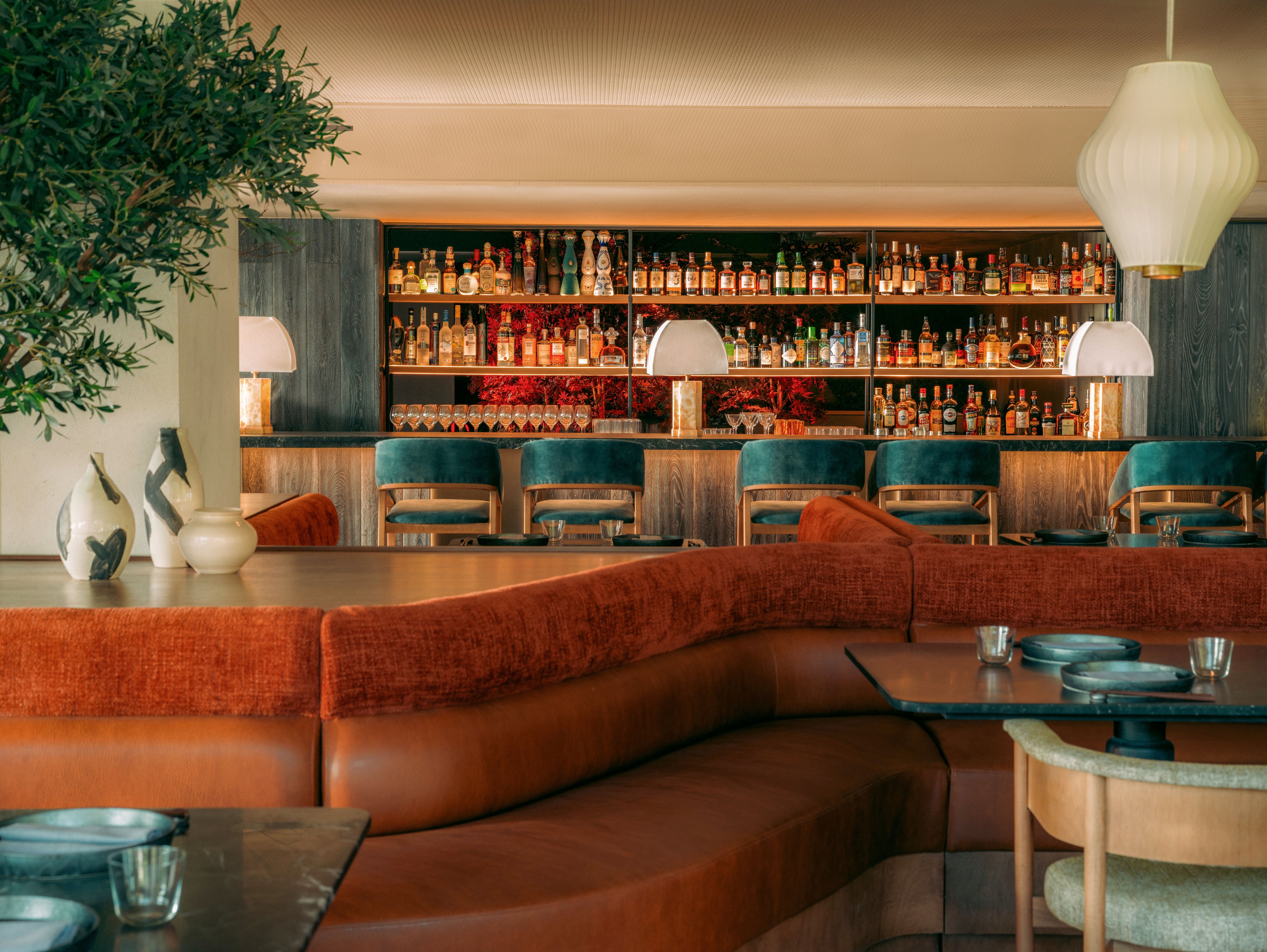 A contemporary bar lounge features a long, curved upholstered banquette in warm rust tones facing sleek dark tables. In the background, a well-stocked bar with rows of bottles is framed by teal bar stools and soft, glowing table lamps. Pendant lighting and sculptural vases add a refined, mid-century modern touch to the space. The overall atmosphere is cozy yet sophisticated, ideal for evening socializing or relaxed cocktails.
