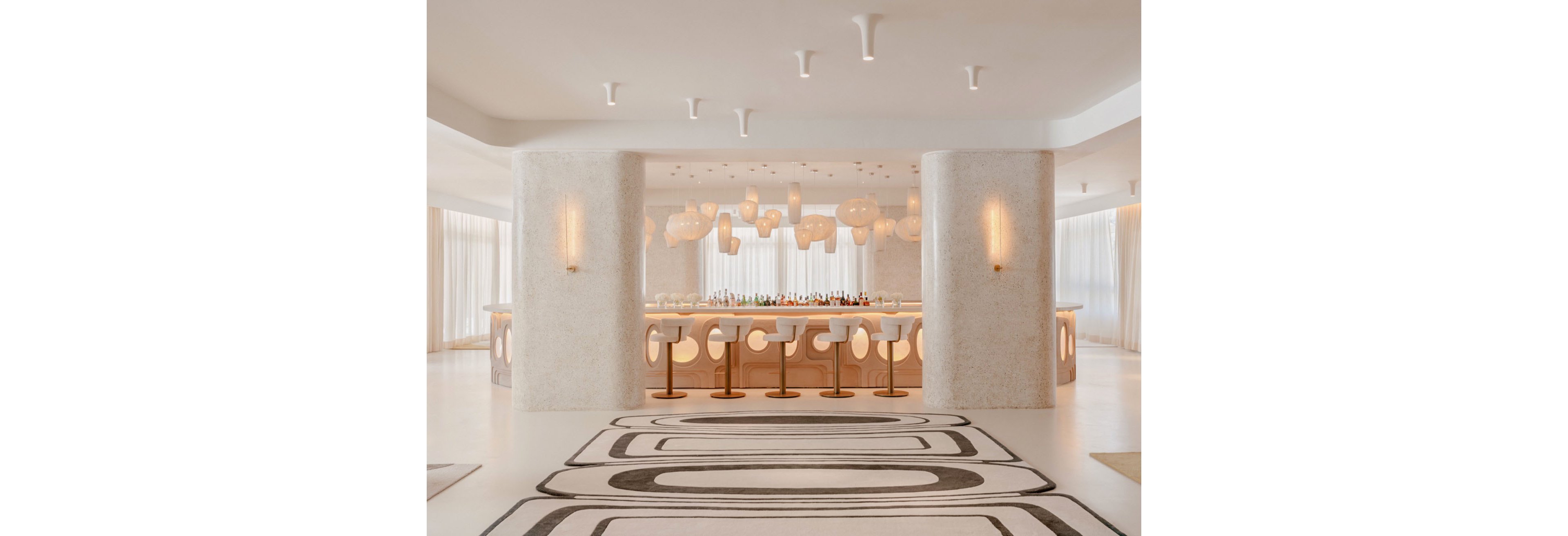A contemporary bar interior features a central counter framed by two large textured columns. Curved white bar stools line the counter, facing an array of colorful bottles and glassware. Organic, sculptural pendant lights hang from the ceiling, casting a warm glow over the space. Soft neutral tones, flowing curtains, and a bold geometric floor pattern create a refined, modern atmosphere.