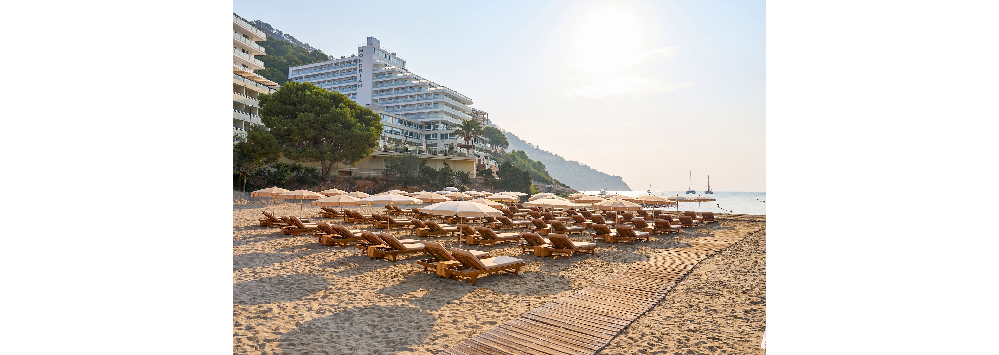 A wide sandy beach is lined with rows of wooden sun loungers and beige umbrellas, all currently empty. A wooden boardwalk leads across the sand toward the calm sea, which stretches into a hazy horizon. Modern multi-story resort buildings rise beside the shoreline, framed by green trees and distant hills. The soft morning light and gentle colors create a peaceful, tranquil vacation atmosphere.