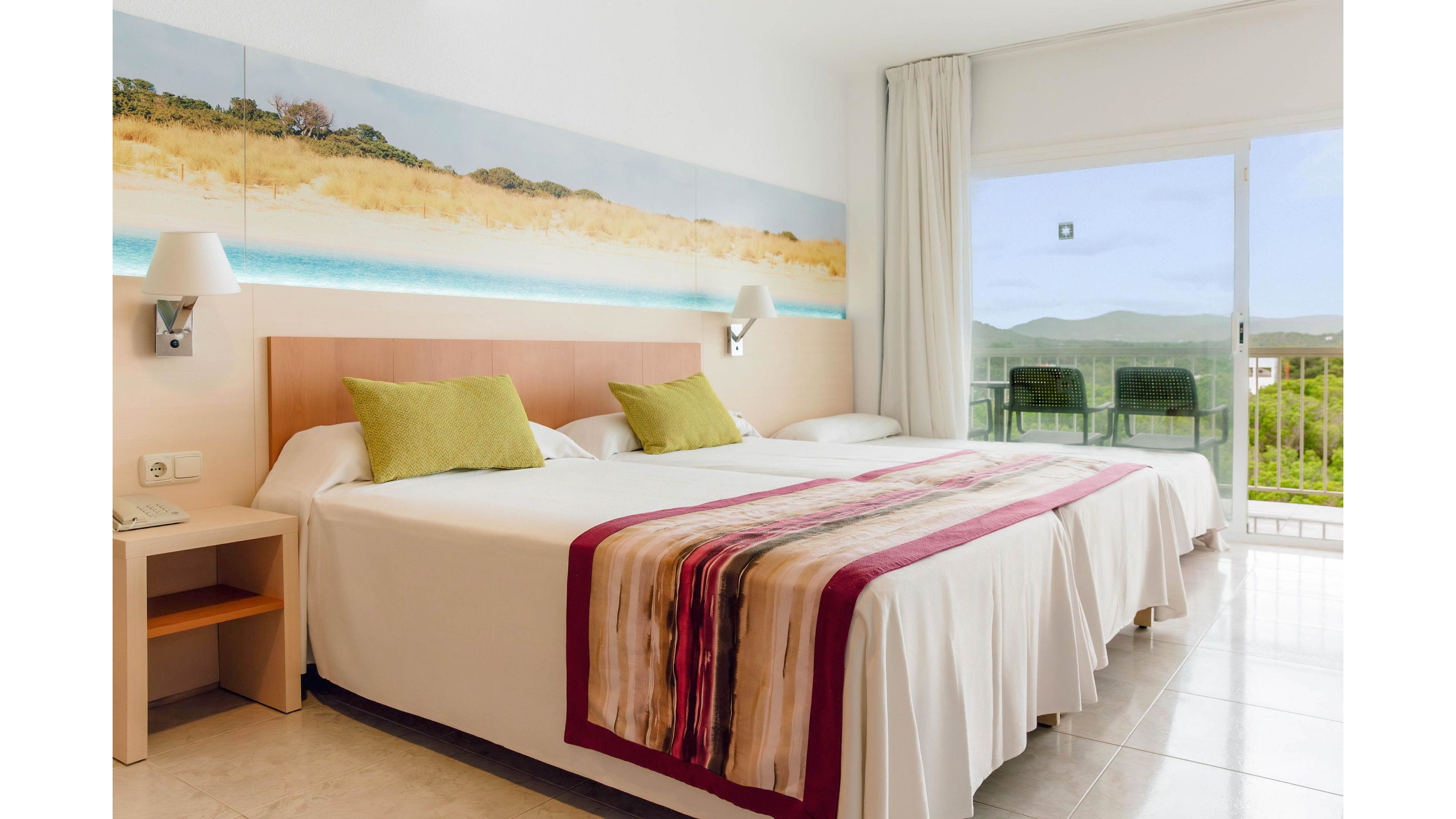 Modern hotel room featuring two twin beds pushed together with white linens and colorful striped runner. Green accent pillows and a large beach landscape artwork add a fresh, coastal feel to the space. A bedside table with wall-mounted lamps sits beside the bed, and light curtains frame a large glass door. The balcony outside offers chairs and a scenic view of green hills under a clear sky.