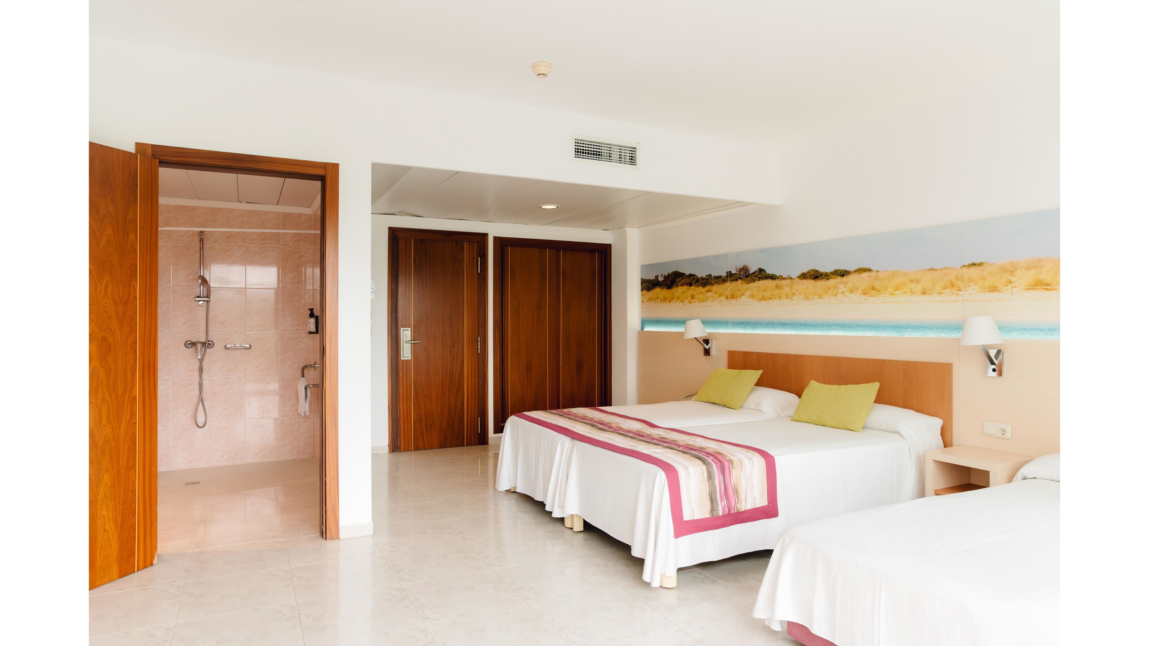 A bright, modern hotel room features two neatly made twin beds with white linens and colorful accent runners and pillows. Wooden doors lead to a bathroom with a walk-in shower visible on the left side. A long beach-themed mural decorates the wall above the beds, adding a coastal touch to the interior. The space is clean, minimal, and well-lit, suggesting a comfortable vacation stay.