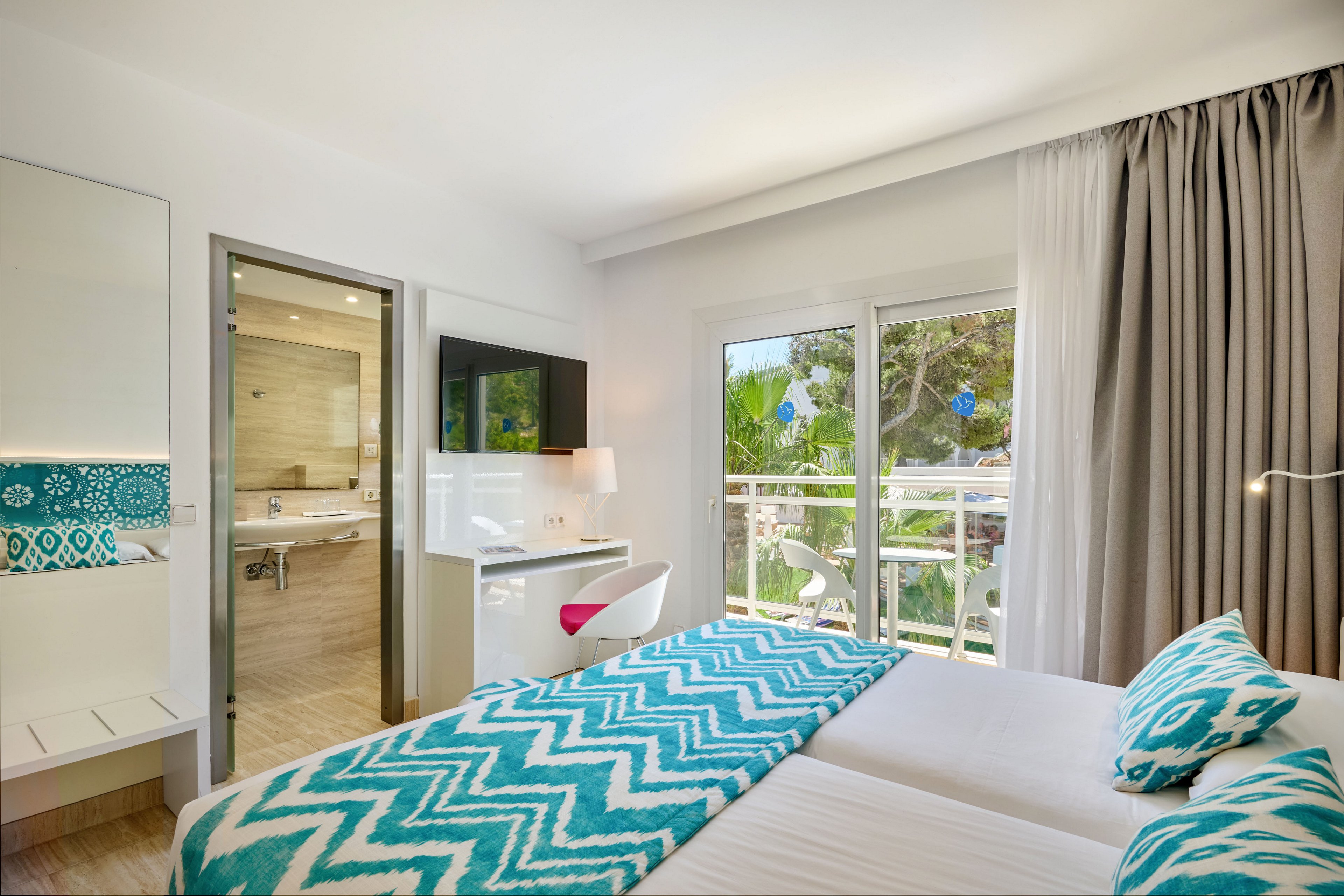 Contemporary hotel room featuring two twin beds with turquoise zigzag patterned throws and pillows. Large glass doors open to a balcony overlooking lush green trees and outdoor scenery. A modern bathroom with a wide vanity mirror and walk-in shower is visible through an open doorway. Neutral walls and curtains contrast with the vibrant teal accents, creating a fresh and inviting atmosphere.