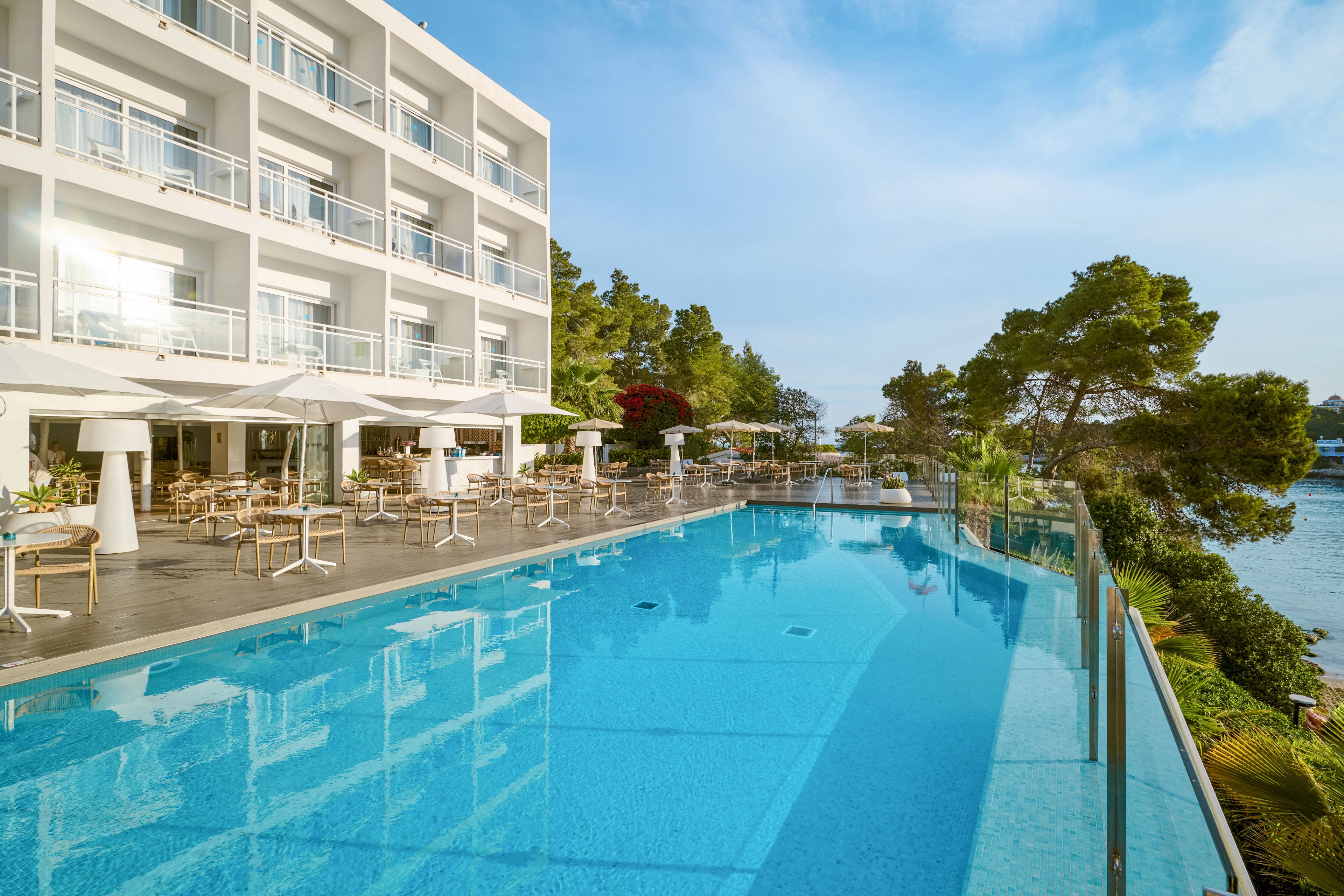 A contemporary multi-story hotel overlooks a large infinity-style swimming pool. Sun loungers and outdoor dining tables with umbrellas line the poolside terrace. Lush green trees and coastal water frame the scene, suggesting a relaxing seaside location. The bright daylight and clear blue sky create a fresh, tranquil vacation atmosphere.