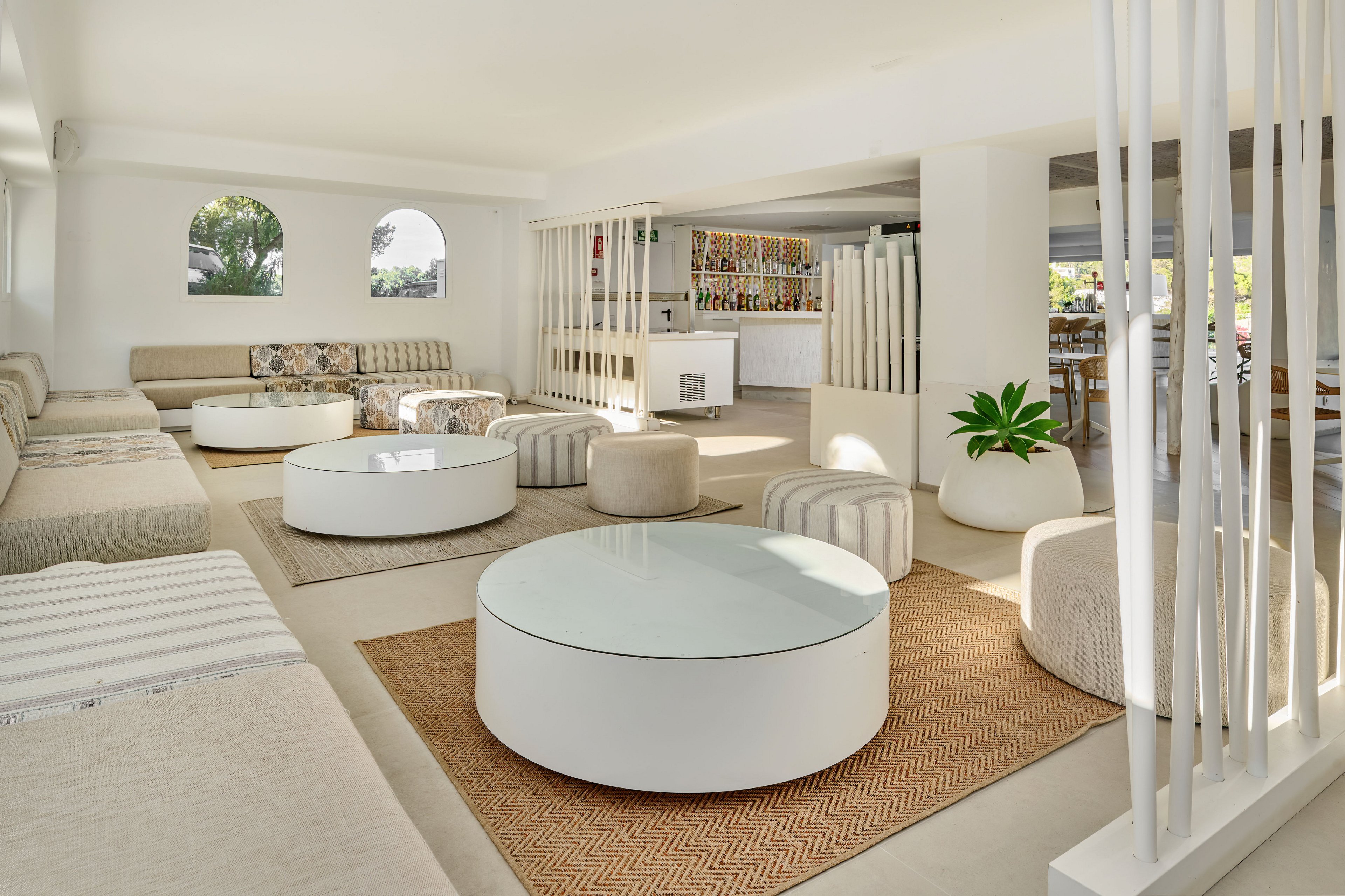 A spacious contemporary lounge features low circular coffee tables and cushioned benches in a bright, open-plan interior. Neutral-toned sofas and poufs are arranged on woven rugs, creating a relaxed social seating area. White walls, columns, and vertical slats add architectural interest, while a green potted plant introduces a touch of nature. Large windows and arched openings allow natural light to flood the room, highlighting the clean, minimalist design.