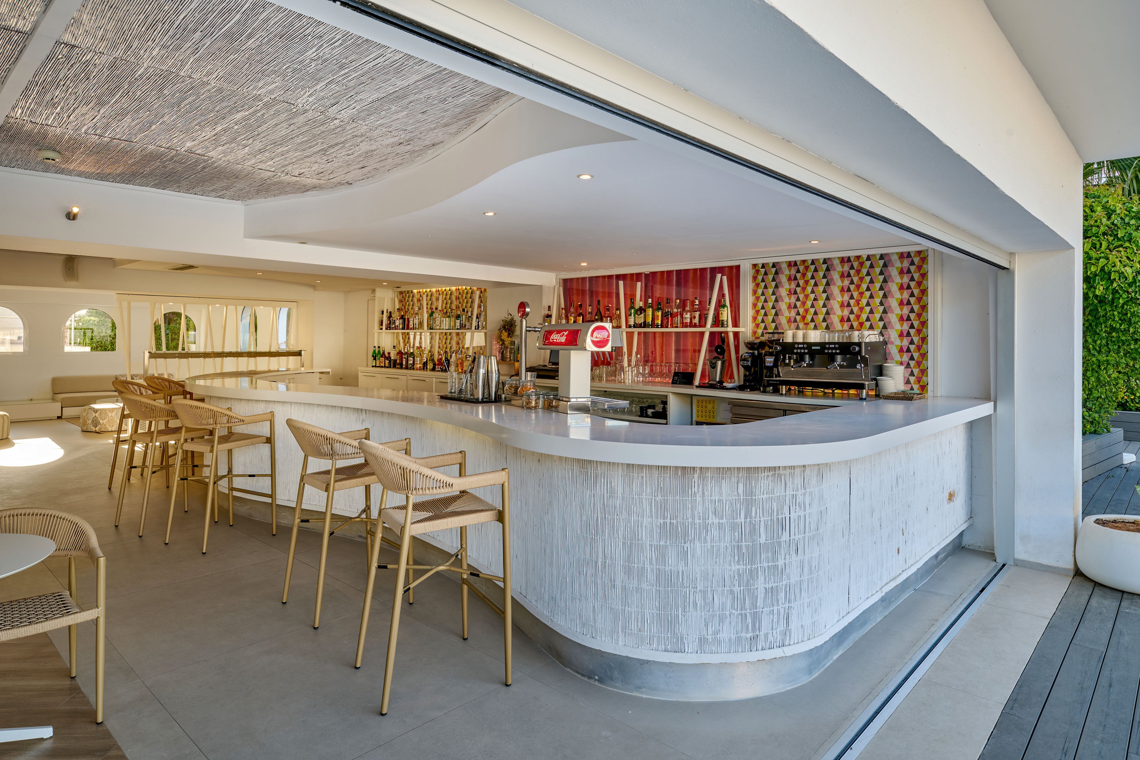 A contemporary open-air bar features a long curved white counter with several light wooden bar stools. The setting appears bright and inviting, with large sliding openings connecting the interior to an outdoor deck. Behind the bar, shelves display bottles and glassware in front of a vibrant multicolored wall. The overall design is clean and modern, emphasizing relaxed socializing in a stylish environment.