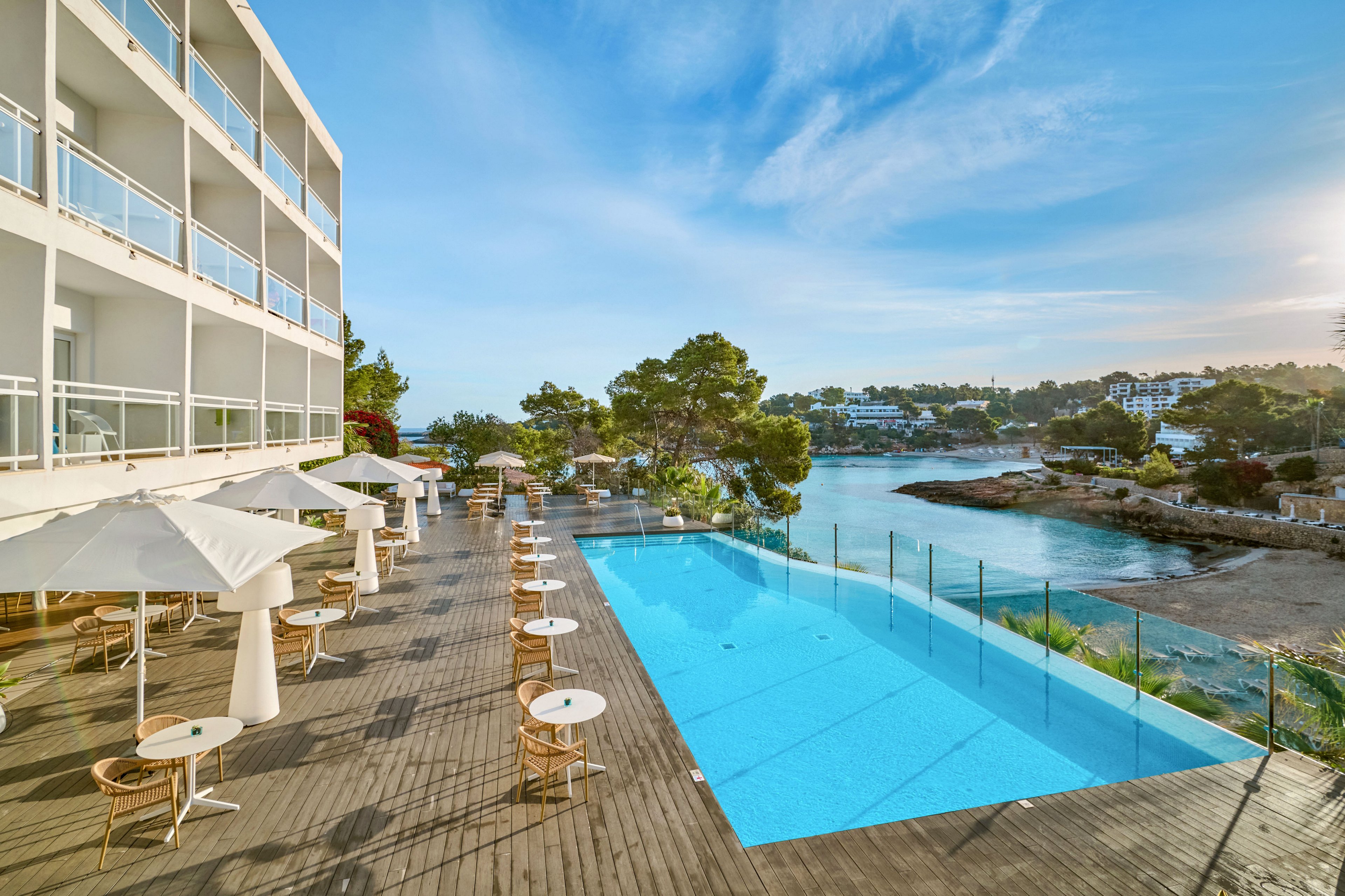 A modern seaside hotel features a spacious wooden terrace overlooking a calm coastal inlet. Sun loungers and white parasols line the deck beside a long, narrow infinity-style swimming pool. Glass railings separate the pool area from the rocky shoreline and blue water beyond. Trees and low-rise buildings are visible across the bay under a clear, sunny sky, creating a relaxed vacation atmosphere.