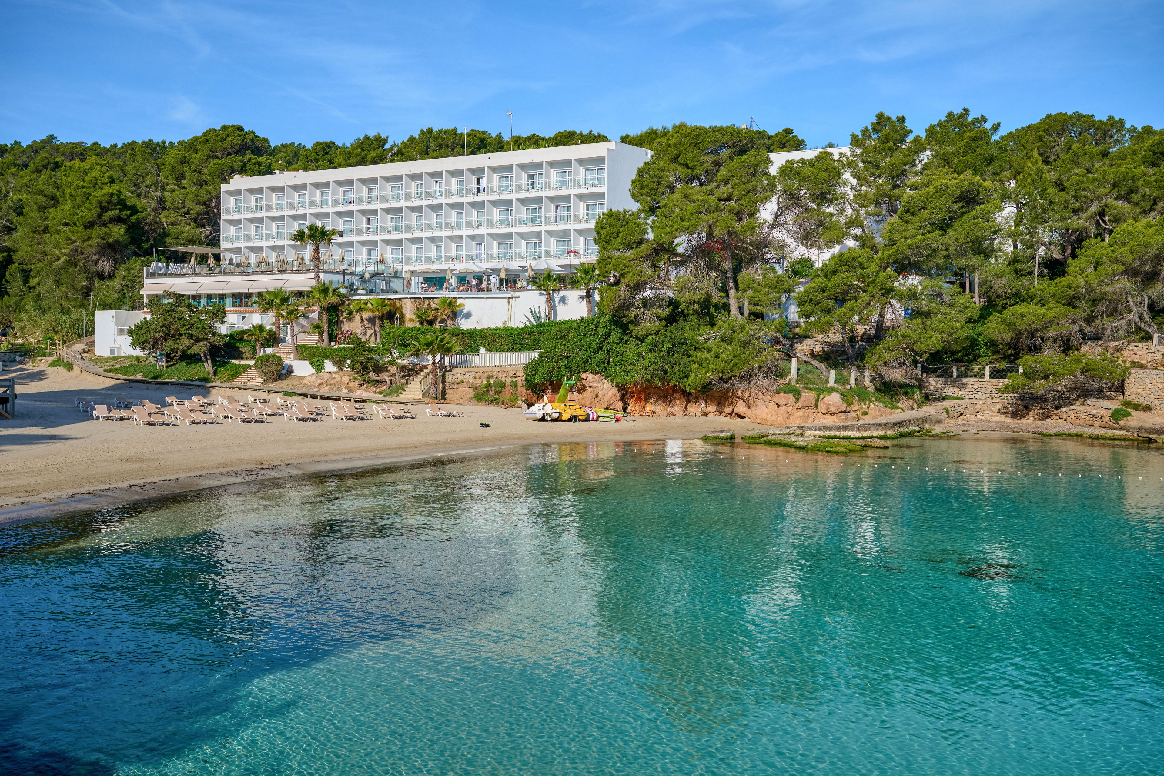 A modern multi-story hotel overlooks a calm turquoise bay. The building is set among dense green pine trees above a sandy beach with sun loungers arranged along the shore. Clear, shallow water in the foreground reflects the blue sky, creating a tranquil coastal scene. The overall atmosphere suggests a relaxing seaside vacation destination.