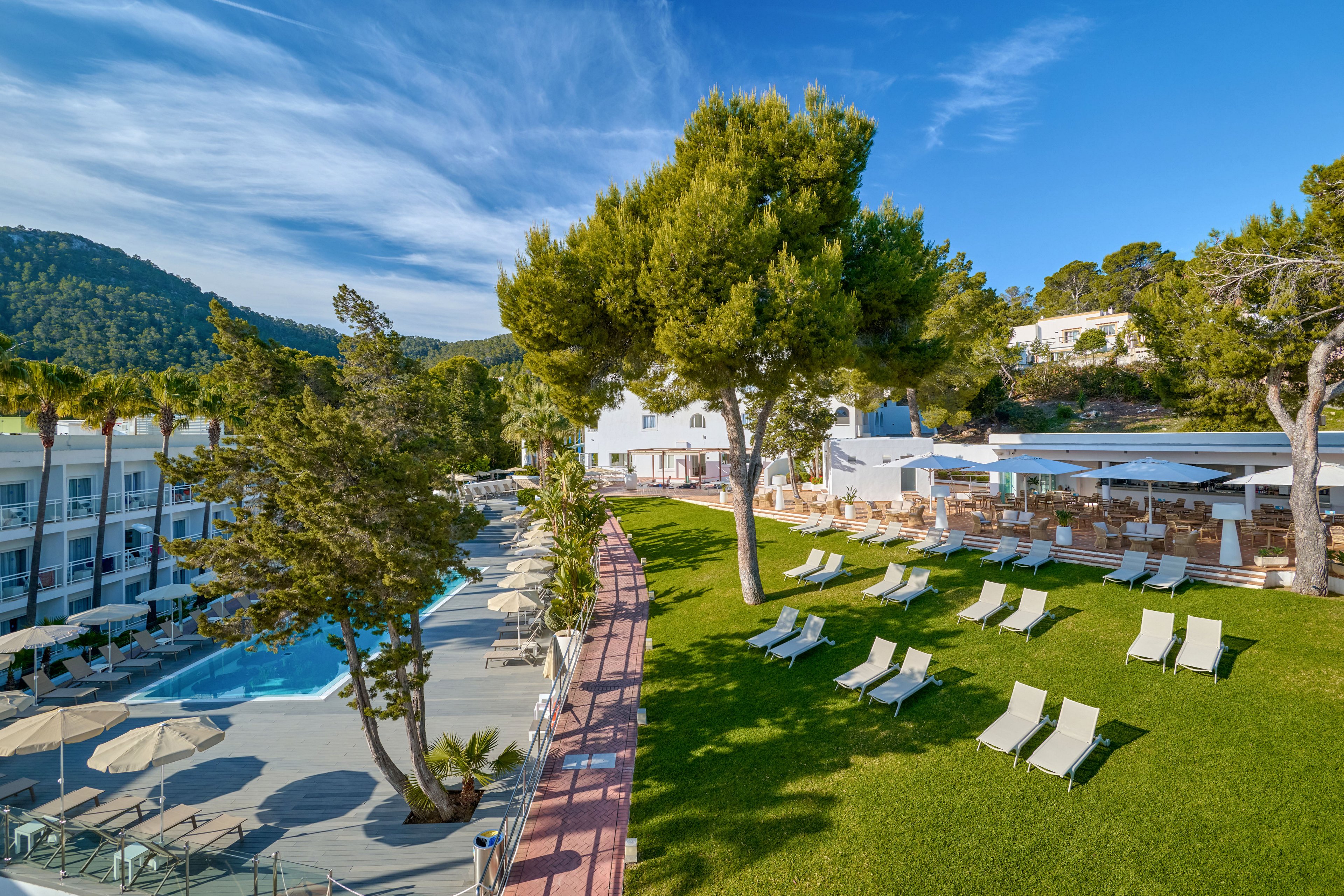 An outdoor resort area features a large green lawn lined with sun loungers and umbrellas beside a swimming pool. Modern white buildings and an open-air terrace with seating are visible in the background. Tall trees and surrounding forested hills frame the scene under a bright blue sky with wispy clouds. The setting suggests a tranquil vacation destination focused on relaxation and leisure.