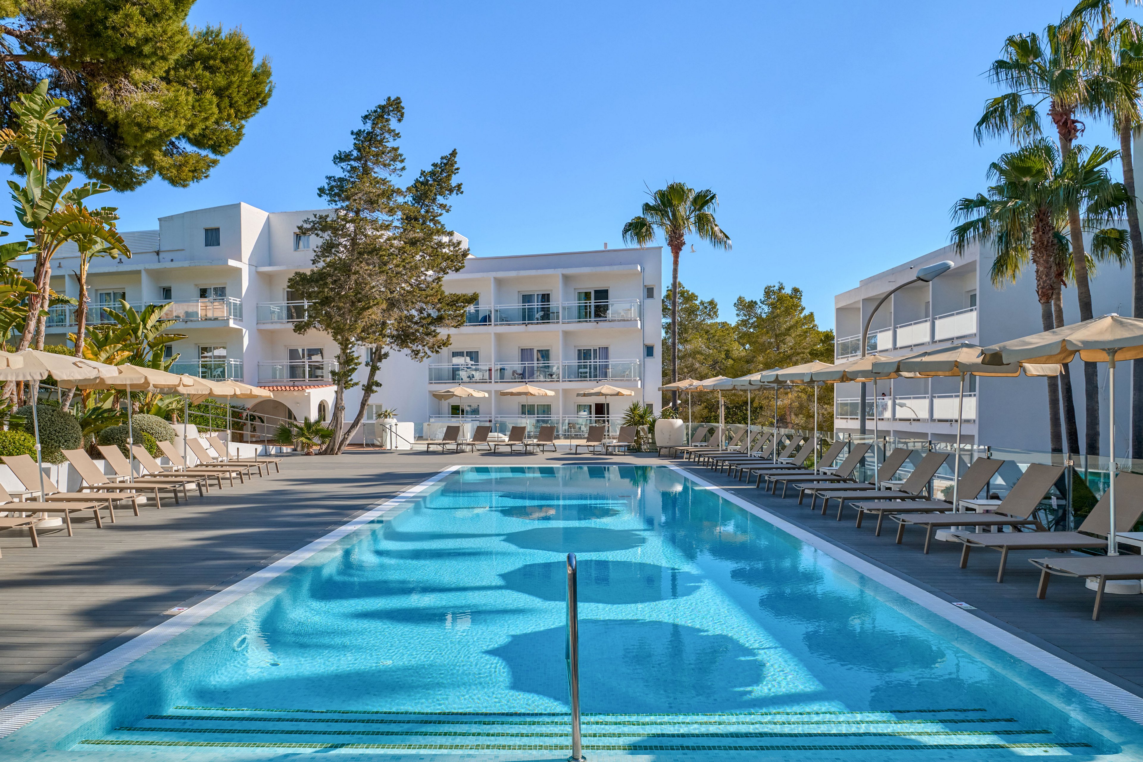 A long rectangular swimming pool stretches between rows of sun loungers and umbrellas at a contemporary resort. White multi-story buildings with balconies frame the pool area, surrounded by palm trees and greenery. The clear blue water reflects the sunny sky, creating a bright and inviting atmosphere. The scene suggests a relaxing vacation setting with ample outdoor seating and modern architecture.