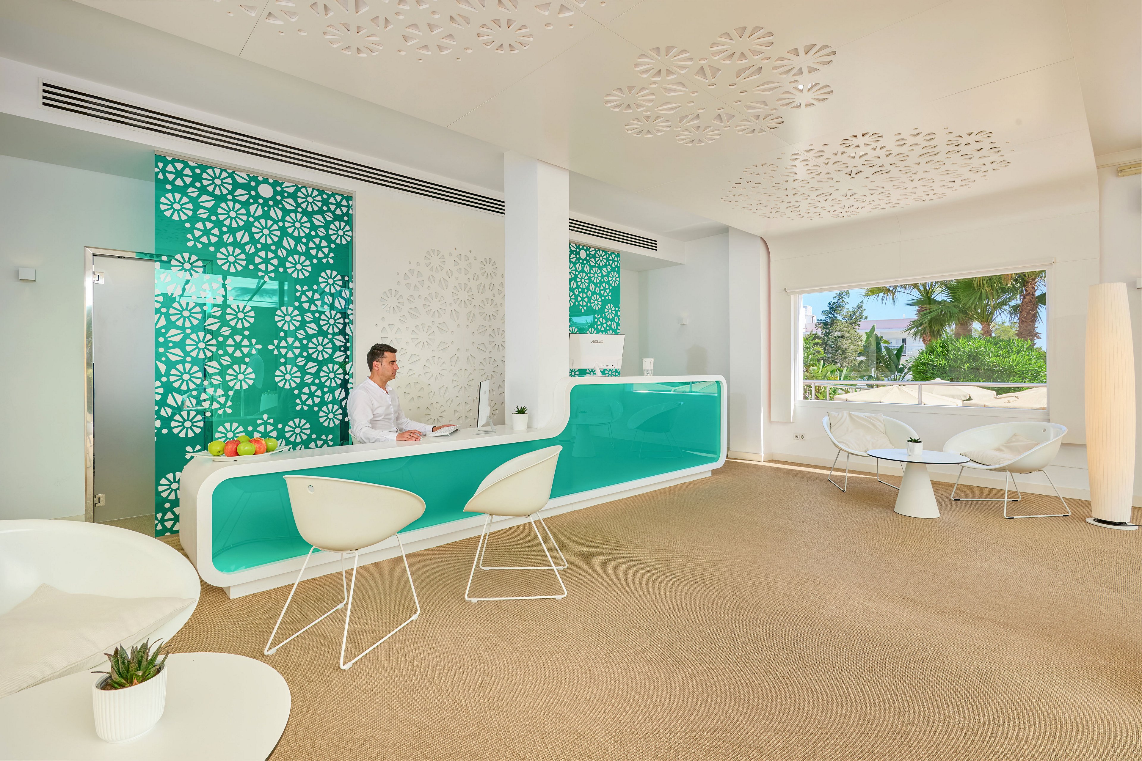A hotel receptionist sits behind a sleek turquoise and white front desk in a bright, contemporary lobby. Decorative wall panels with floral cutout patterns and matching ceiling accents create a cohesive design. Large windows reveal palm trees and outdoor seating, enhancing the tropical atmosphere. Minimalist white chairs and tables complete the clean, modern interior style.