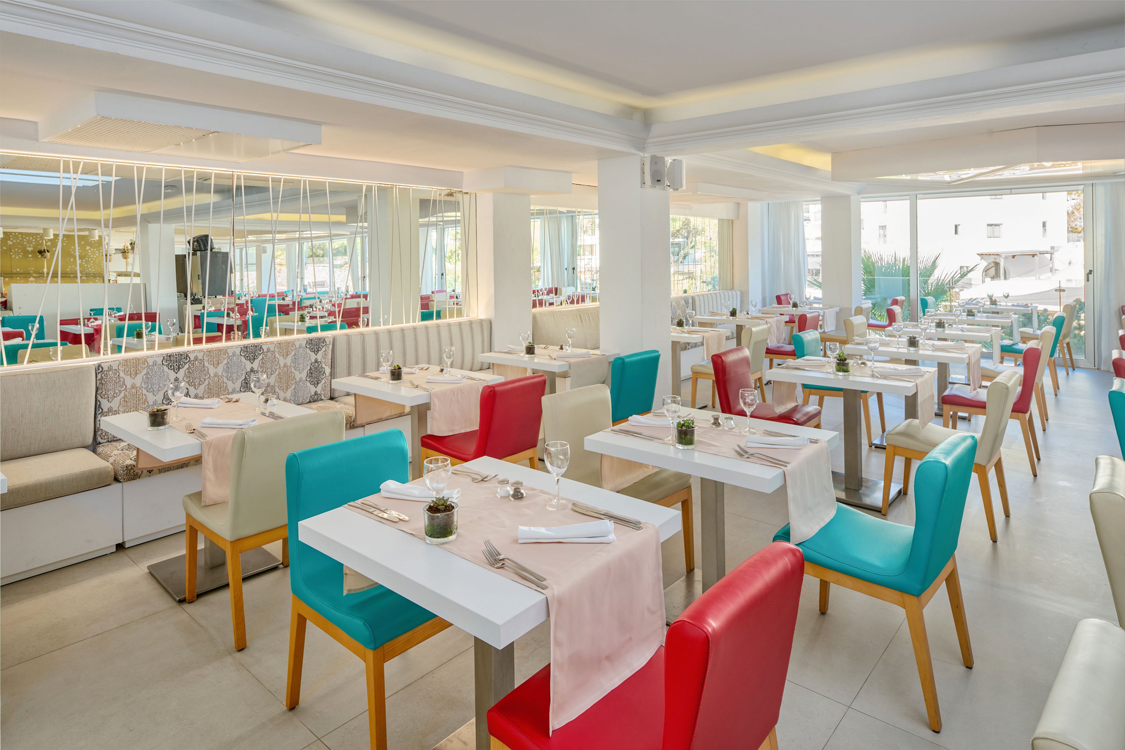 Contemporary restaurant dining area featuring white tables and vibrant turquoise and red chairs. The space is well-lit with large windows and light-colored walls, creating an airy atmosphere. Tables are neatly set with glassware, cutlery, and pale pink table runners, suggesting readiness for guests. Neutral bench seating and decorative elements add comfort and style to the modern interior.