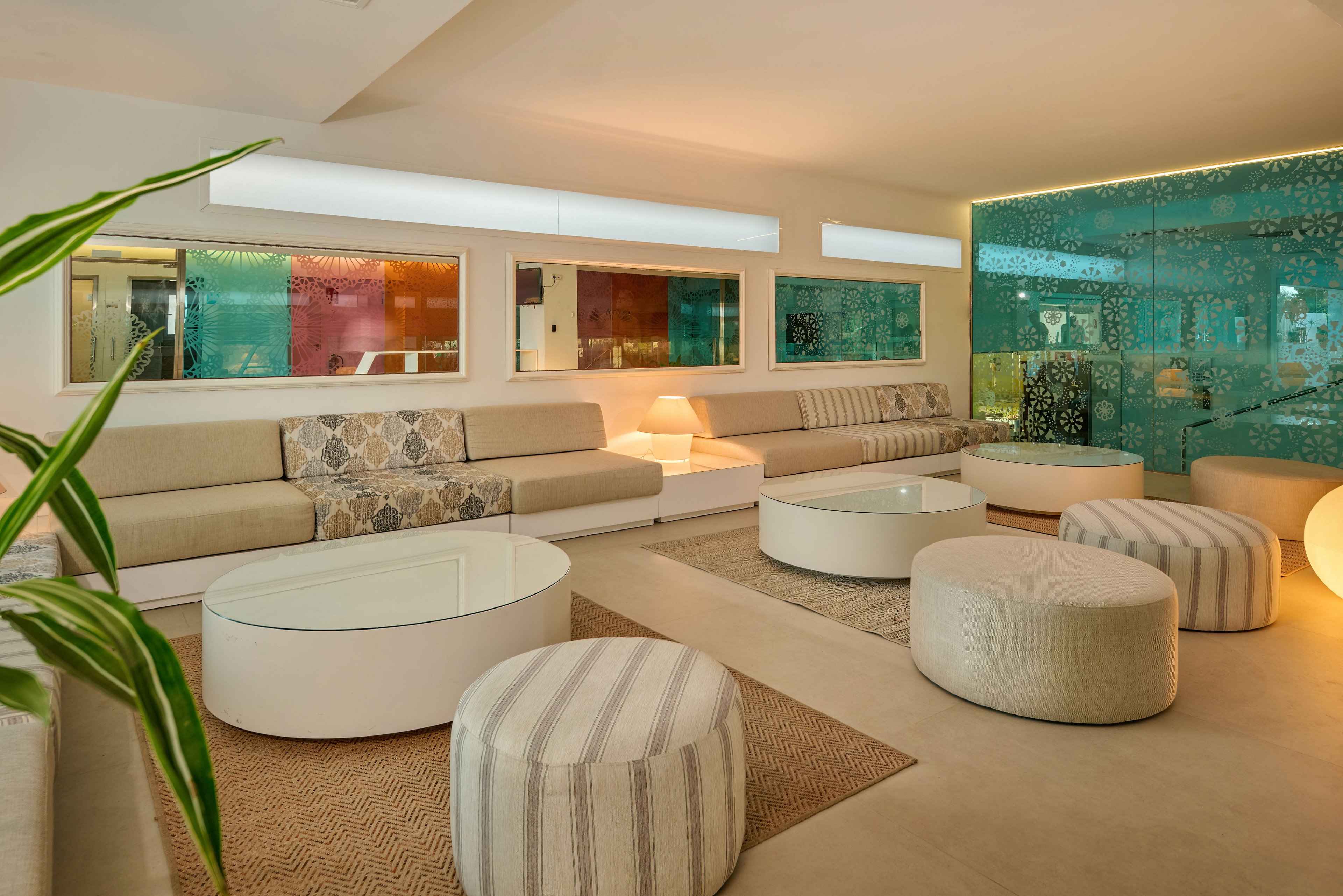 Contemporary lounge interior featuring a long beige sectional sofa and multiple round ottomans. The space includes low circular coffee tables and a textured area rug on a smooth floor. A turquoise decorative wall with patterned cutouts adds visual interest alongside colorful window panels. Soft, warm lighting and neutral tones create a relaxed, inviting atmosphere.