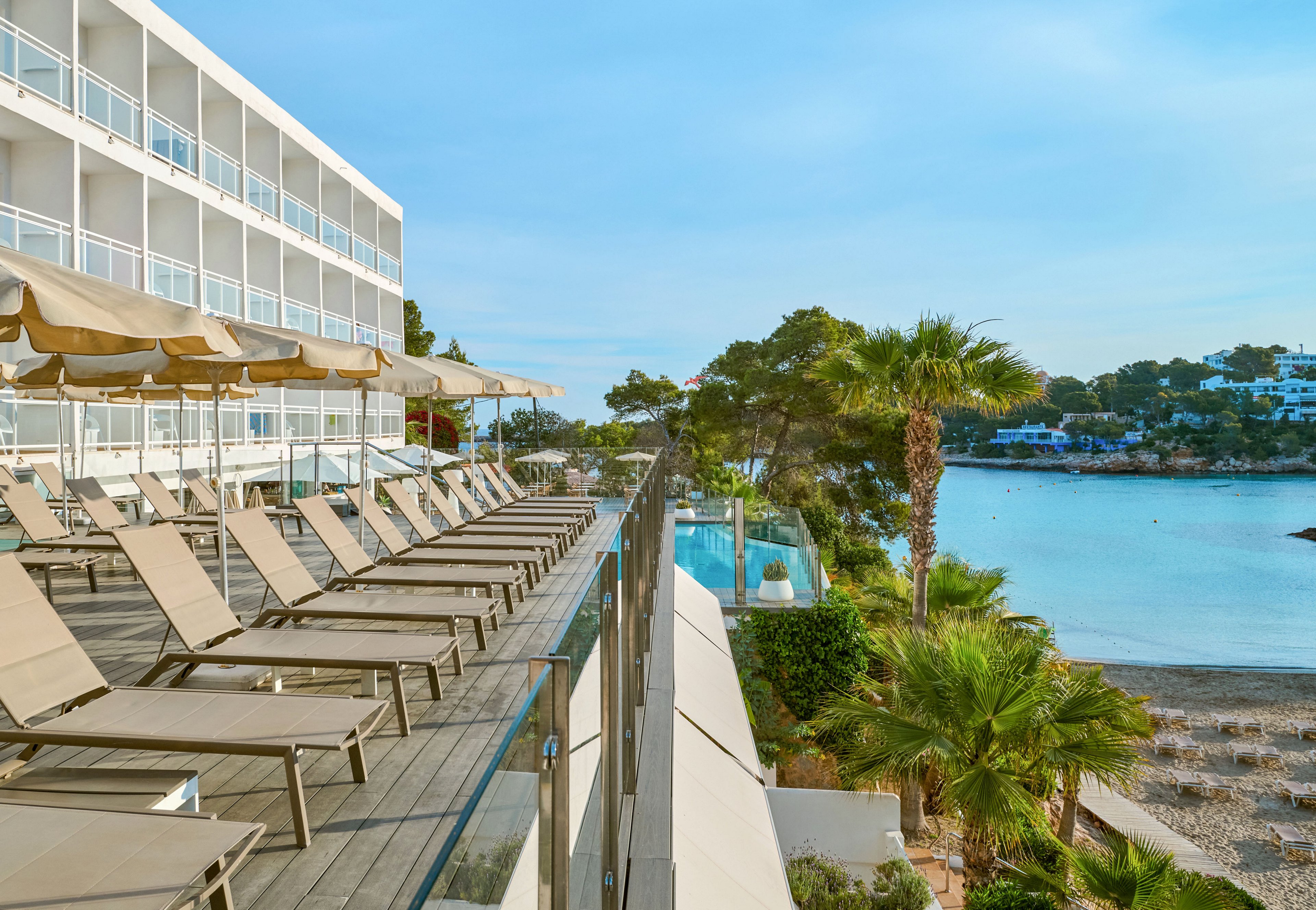 A modern seaside hotel features a long terrace lined with empty sun loungers and large parasols. Glass railings separate the deck from lush palm trees and greenery below. In the background, calm blue water and distant white buildings create a tranquil coastal scene. The bright daylight and clear sky emphasize a relaxed vacation atmosphere.