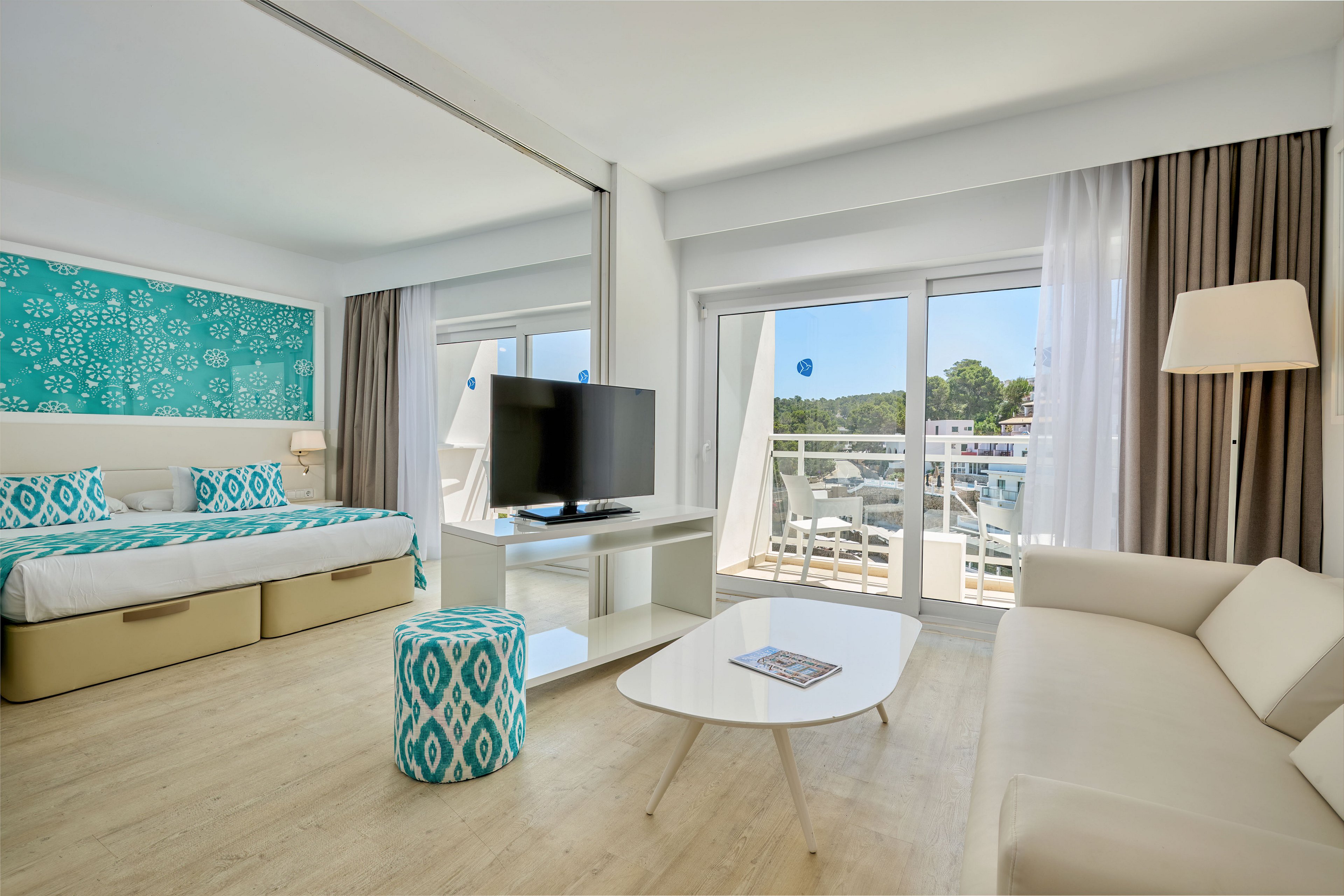 Modern hotel room featuring a double bed with turquoise patterned headboard and cushions. A white sofa and oval coffee table sit opposite a flat-screen television on a stand. Large sliding glass doors open to a balcony with outdoor seating and a scenic view of trees and buildings. Light wood flooring, neutral curtains, and teal accents create a fresh, airy atmosphere.