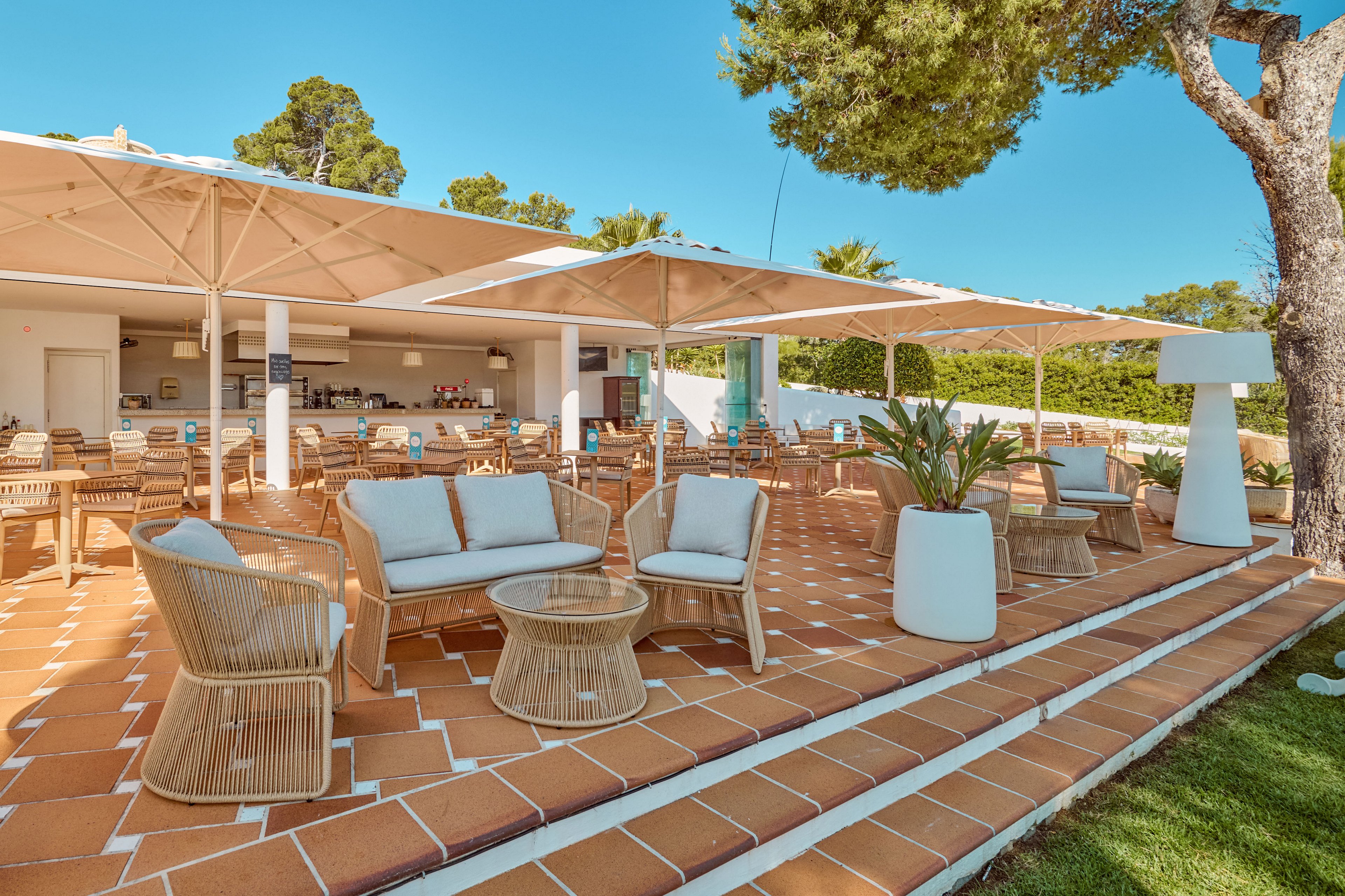 Open-air café terrace with wicker chairs and cushioned sofas arranged on terracotta-tiled steps. Large white umbrellas provide shade over the seating area, leading back to a minimalist bar counter. Potted plants and trees add greenery around the bright, modern patio. The scene suggests a relaxed, resort-style environment under clear blue skies.