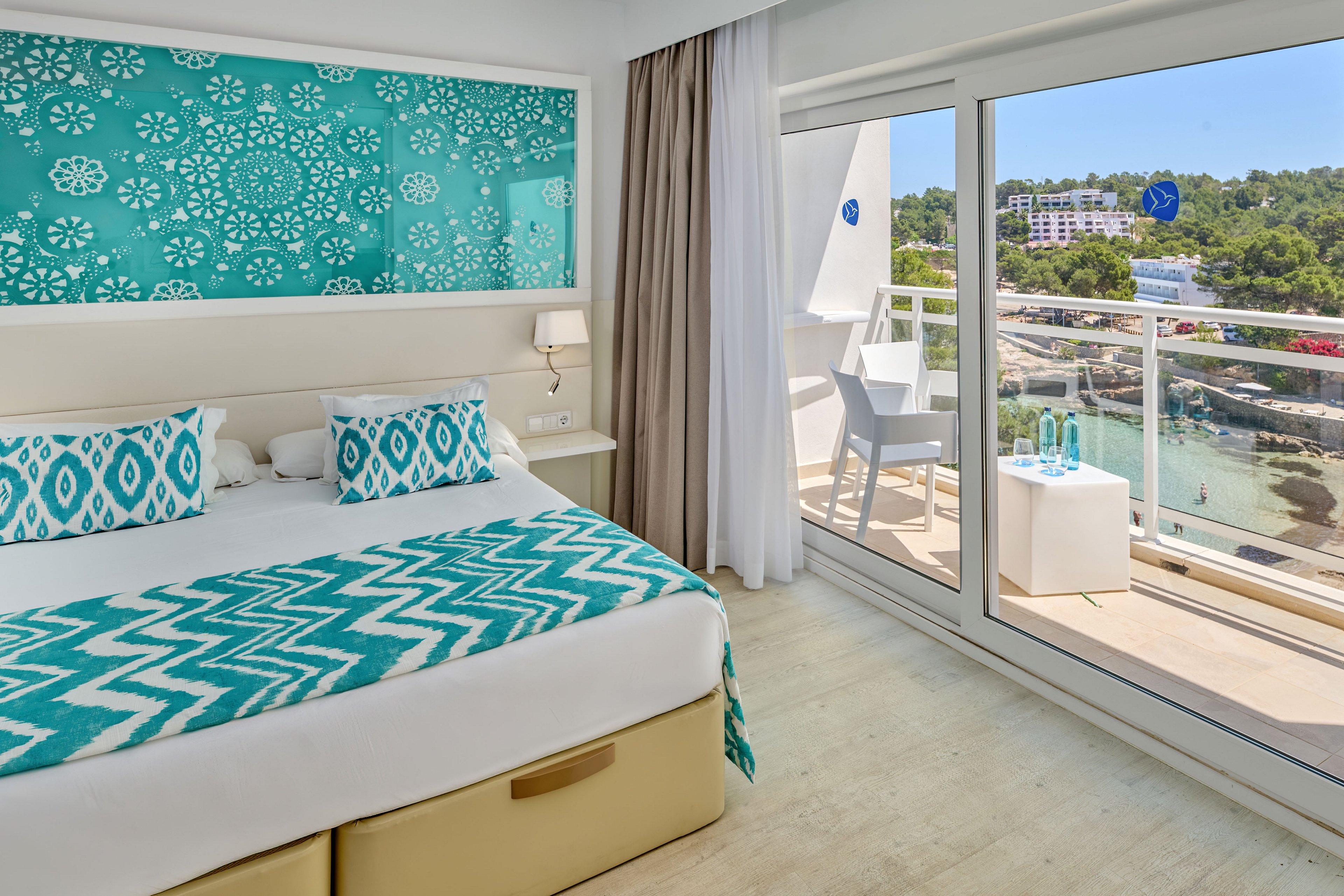 A modern hotel room features a neatly made double bed with turquoise patterned pillows and throw blanket. A large decorative panel with white floral motifs on a teal background hangs above the bed, complementing the light wood floor and neutral curtains. Sliding glass doors open to a balcony with white furniture, two bottles of water, and glasses, overlooking a scenic coastal landscape with trees and buildings. The overall atmosphere is airy, bright, and inviting, emphasizing relaxation and vacation comfort.