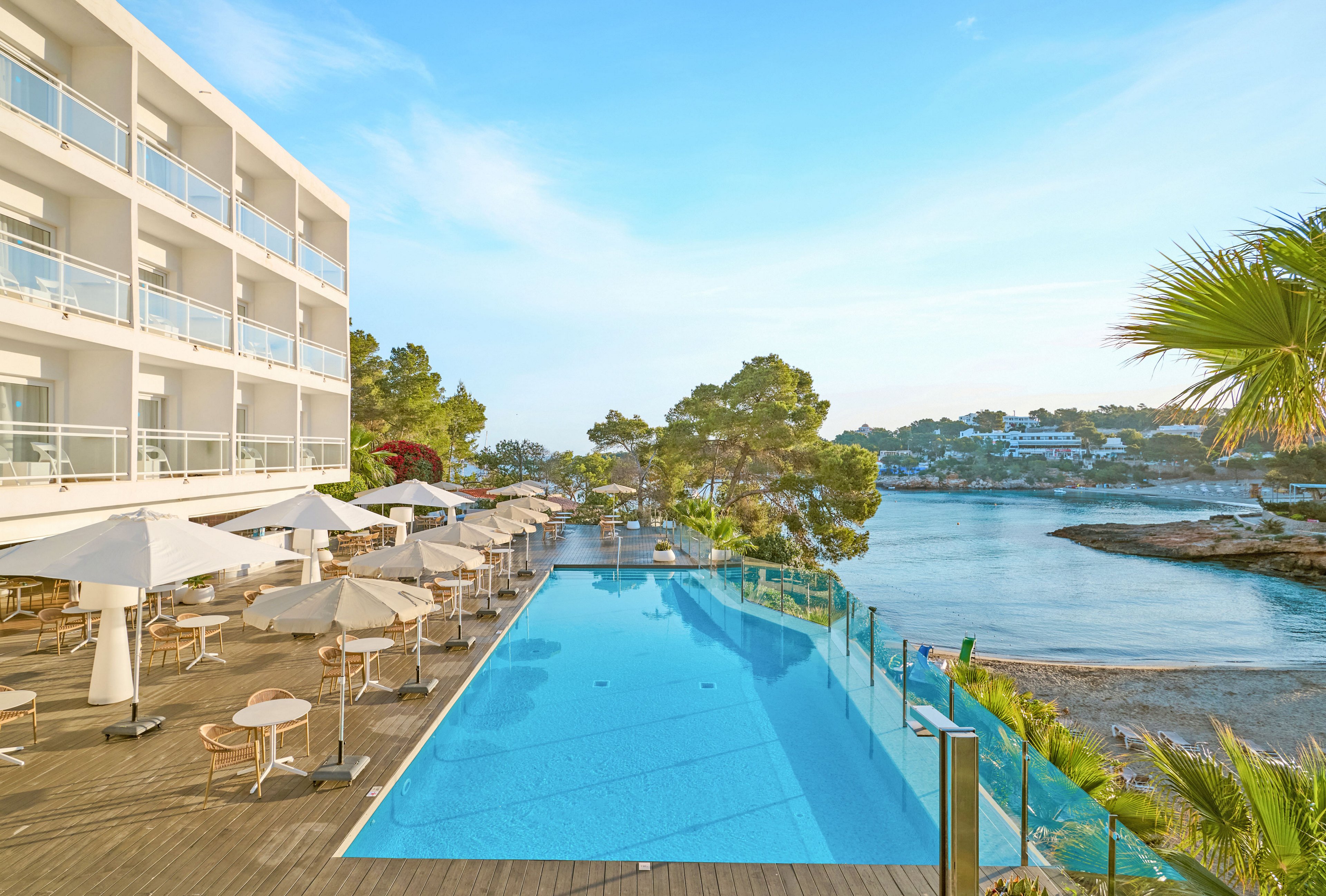 A contemporary multi-story hotel overlooks a long infinity pool beside a calm coastal inlet. Sun loungers and umbrellas line the wooden deck, creating a relaxed vacation atmosphere. Clear blue water in the pool reflects the bright sky, while glass railings frame the view toward rocky shoreline and trees. The scene suggests a tranquil resort destination ideal for leisure and waterfront relaxation.
