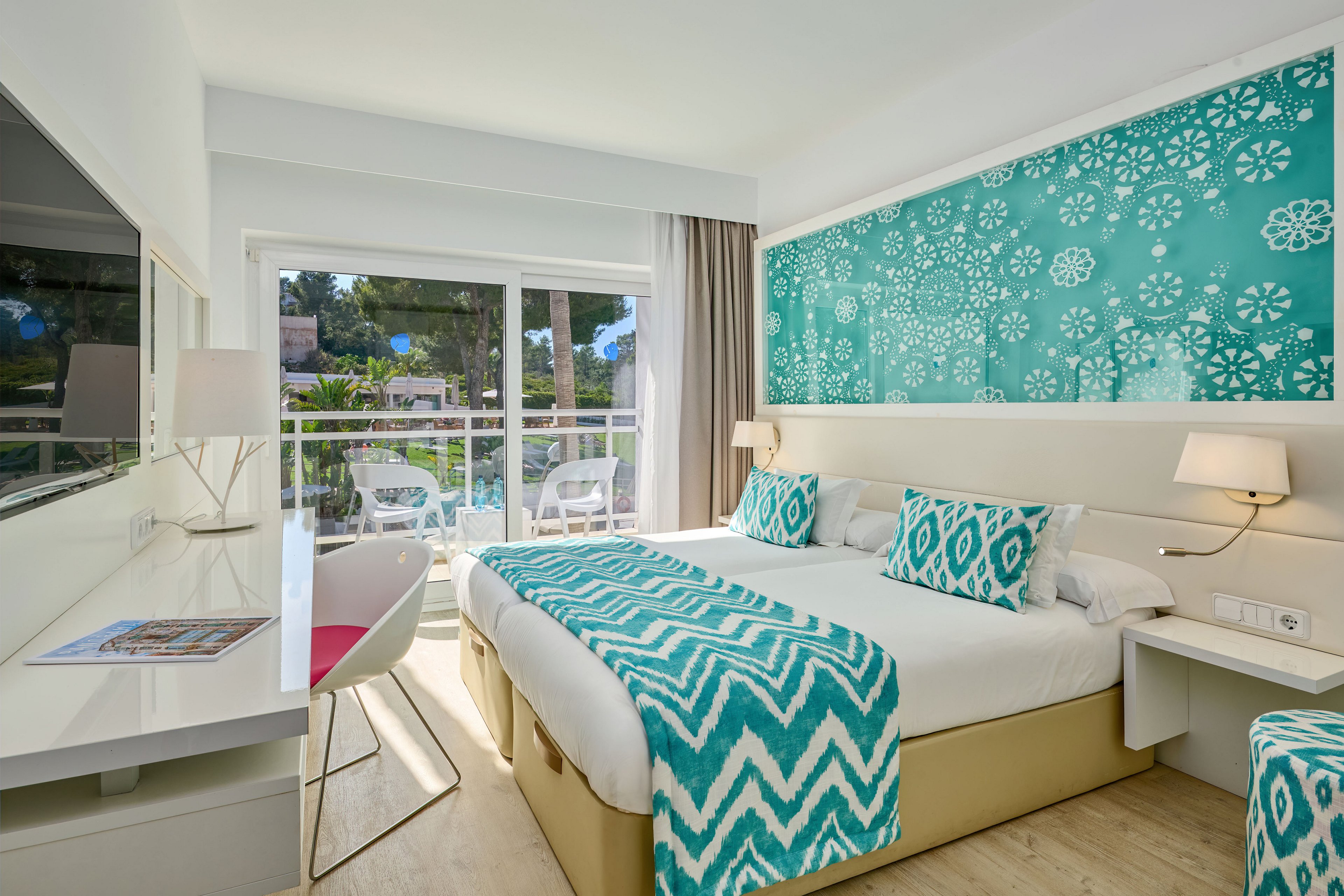 Contemporary hotel bedroom featuring a large double bed with turquoise patterned pillows and throw. The room is decorated with a bold teal accent wall displaying white geometric and floral motifs. A sleek white desk and chair sit opposite the bed, near a large mirror and wall-mounted television. Floor-to-ceiling glass doors open to a balcony with outdoor seating and views of trees and neighboring buildings, creating a light-filled, relaxing atmosphere.