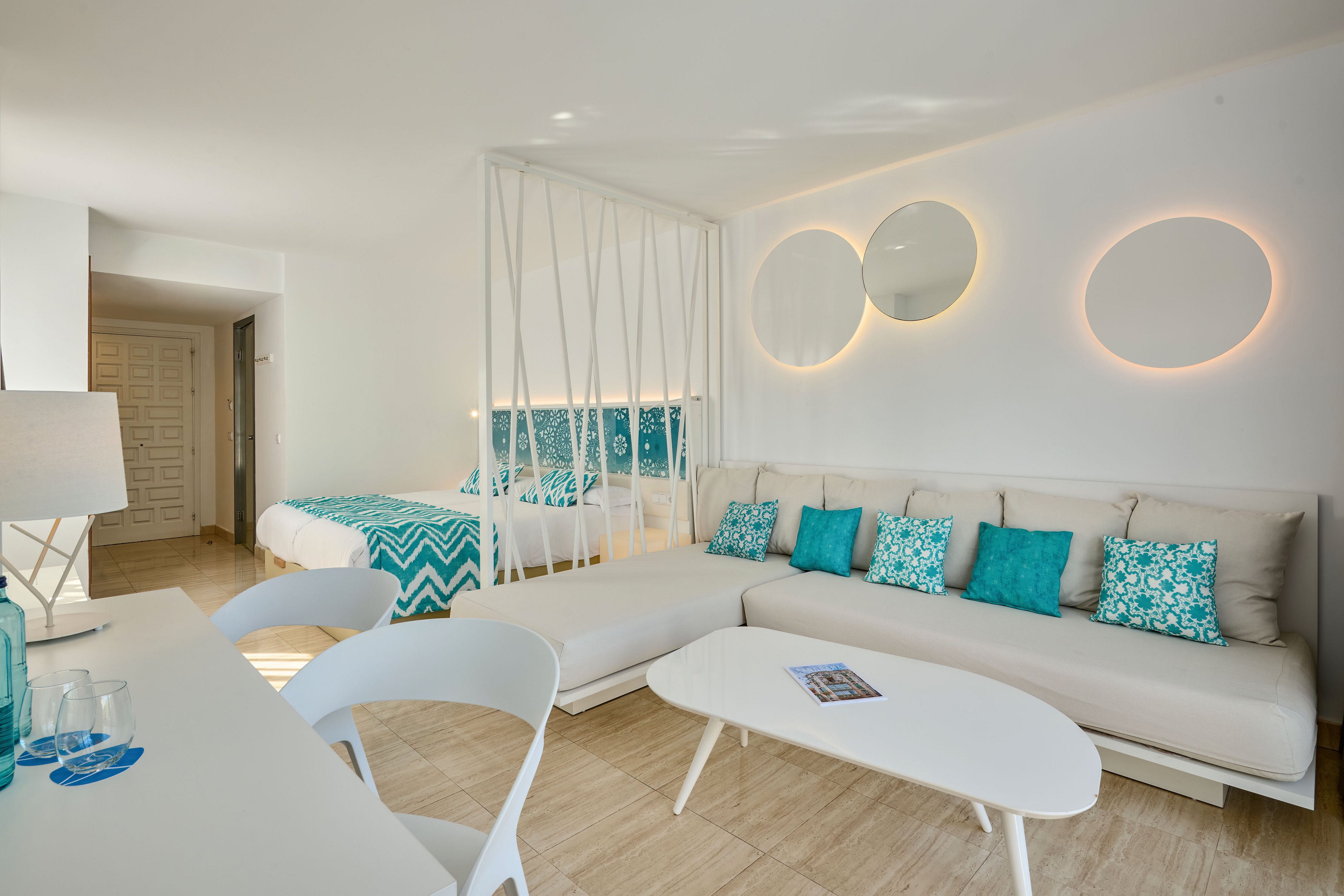 Contemporary hotel room interior featuring a spacious white sofa with teal decorative pillows. A bed with a teal and white chevron-patterned throw is separated by a minimalist white partition. Circular wall lights and mirrors add a stylish focal point to the clean, neutral walls. A white coffee table and desk area with chairs complete the airy, beach-inspired design.