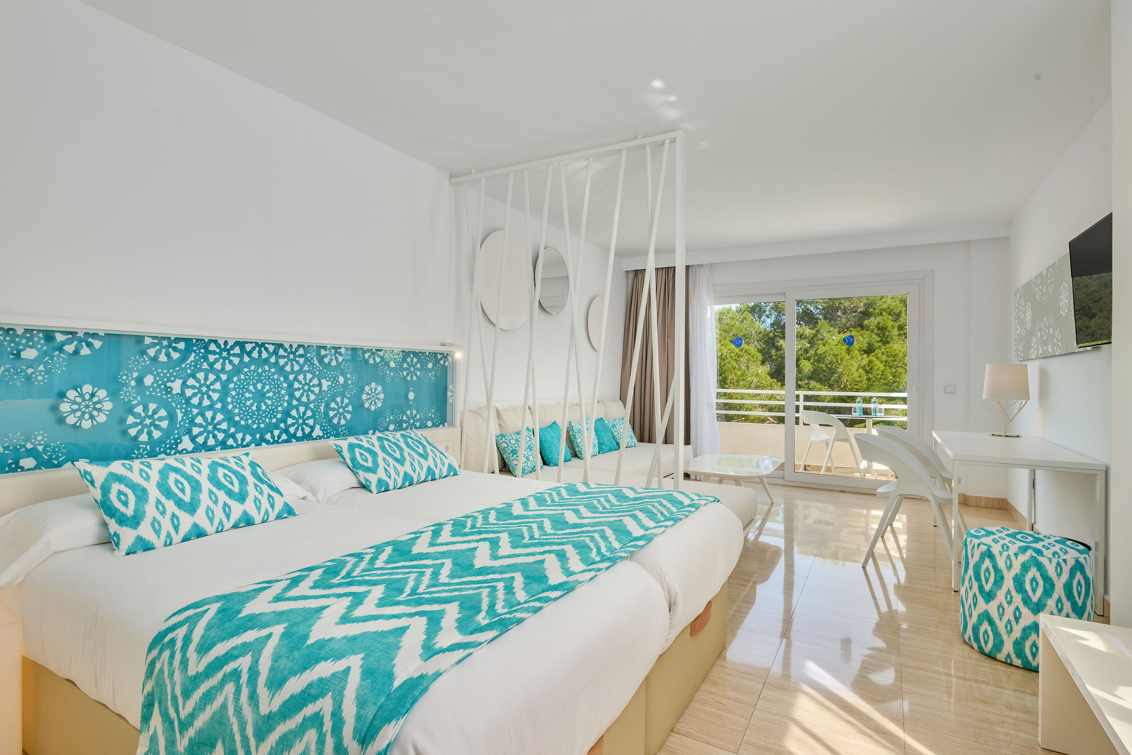 A spacious, contemporary hotel room features two large beds with turquoise patterned throws and pillows against a decorative blue headboard. The interior is predominantly white, creating a clean and airy atmosphere complemented by natural light from a large glass door. A small seating area with white chairs and table sits near the balcony, which overlooks green trees outside. Additional accents include a wall-mounted television, a white desk, and a matching turquoise patterned stool.