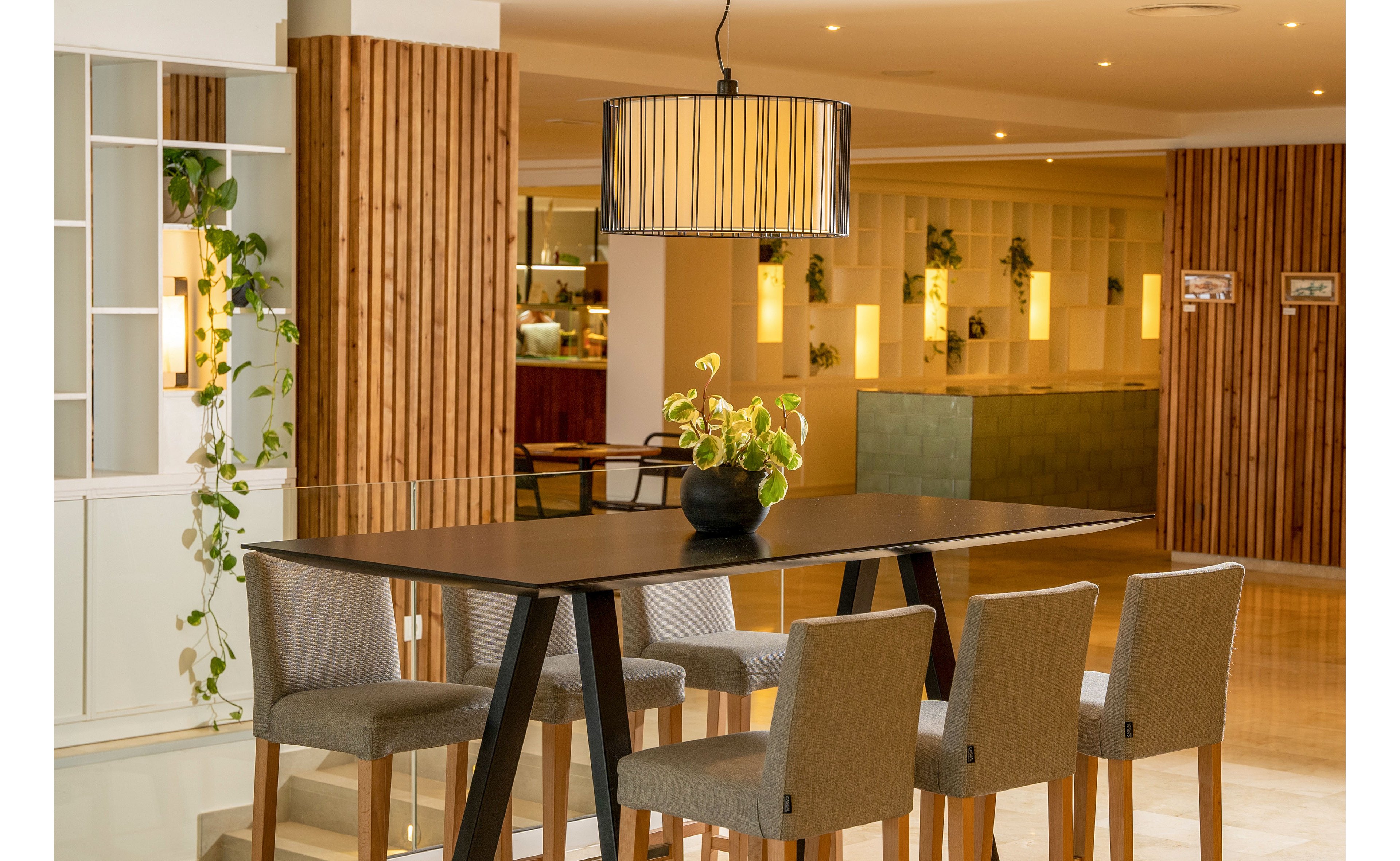 A sleek dark wooden dining table with six upholstered chairs stands in the center of a contemporary interior. A large cylindrical pendant light hangs above the table, illuminating a potted plant centerpiece. The background features wooden slat wall panels, built-in shelving with trailing plants, and softly lit niches. The overall setting suggests a hotel lobby or communal dining area with a warm, inviting atmosphere.