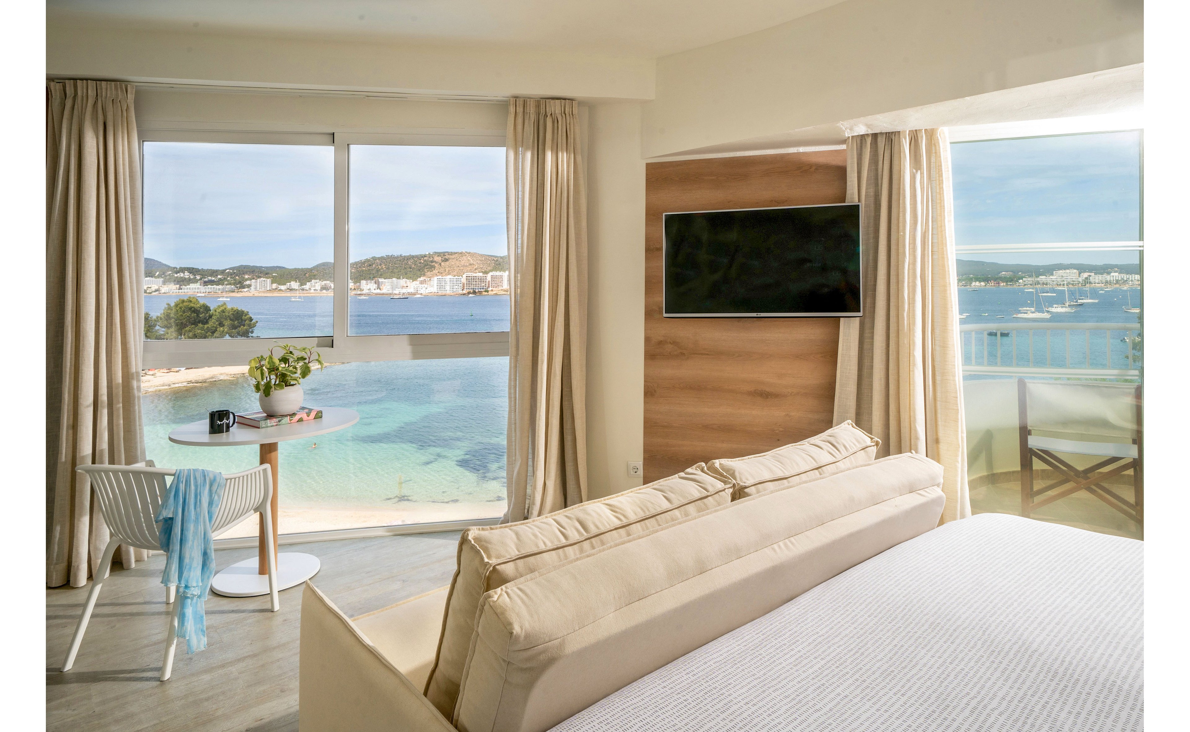 A bright, modern hotel room features a large bed and a beige sofa facing a wall-mounted flat-screen TV. Floor-to-ceiling windows reveal a clear view of a calm turquoise bay and distant shoreline buildings. A small round table with a potted plant and a chair draped with a light blue towel sits near the window. Sunlight fills the space, highlighting neutral tones and creating a serene, coastal atmosphere.