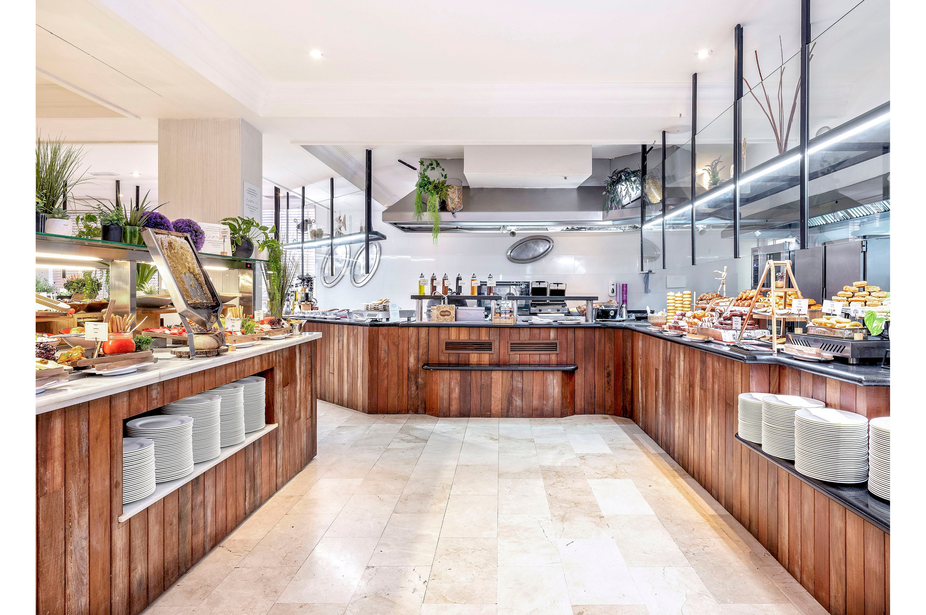 A spacious self-service buffet counter displays a wide variety of foods in a bright, modern dining area. Wooden counters with neatly stacked white plates frame the central serving space. Glass sneeze guards, metal fixtures, and decorative plants add a contemporary touch. The scene is well-lit, emphasizing cleanliness and abundance, with no people present.