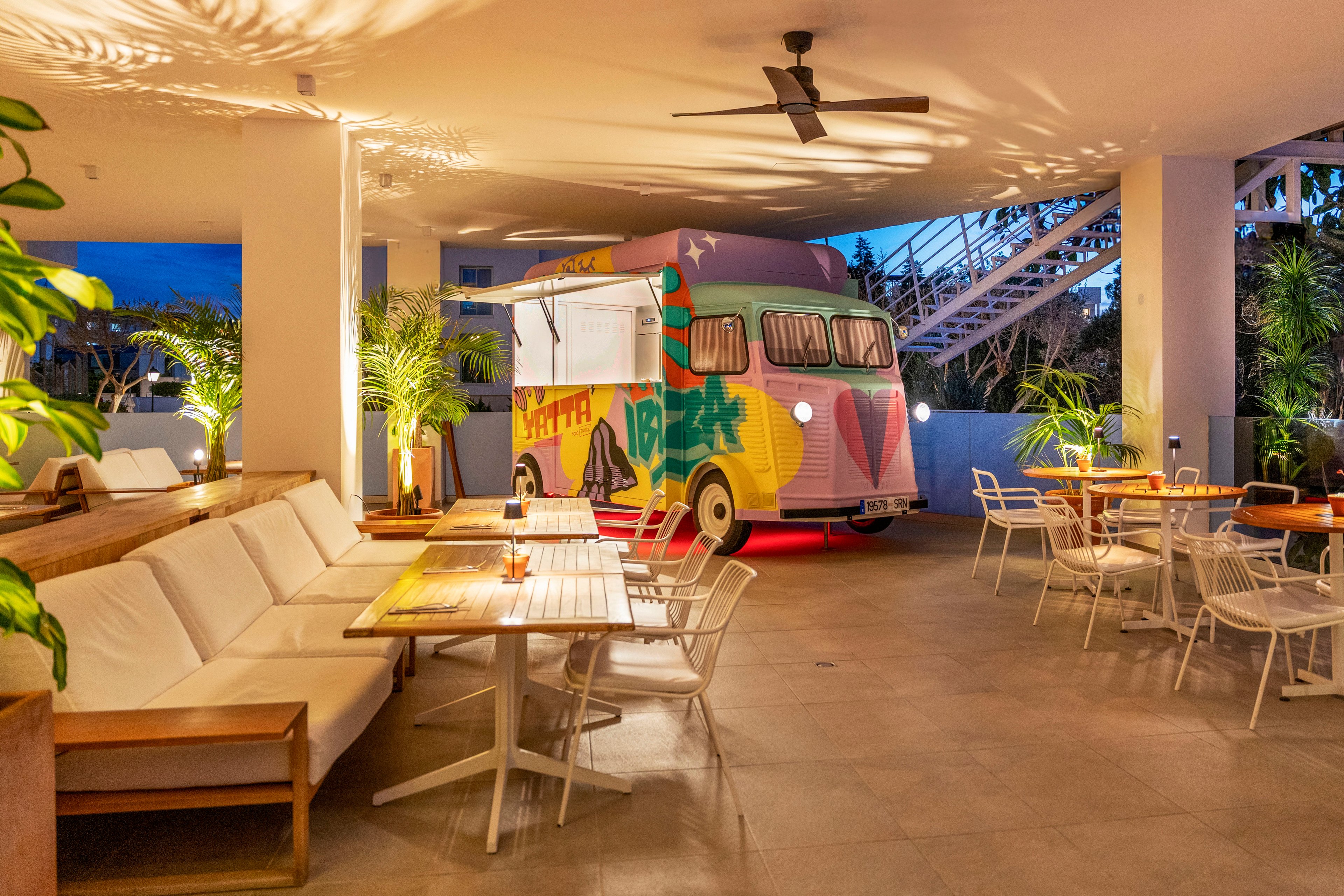 A brightly painted retro food truck is parked inside a modern open-air lounge. Wooden benches with white cushions and white metal chairs surround polished tables, creating a relaxed dining area. Warm ambient lighting and ceiling fans enhance the cozy evening atmosphere. Green potted plants and an outdoor staircase frame the scene, adding a tropical touch.