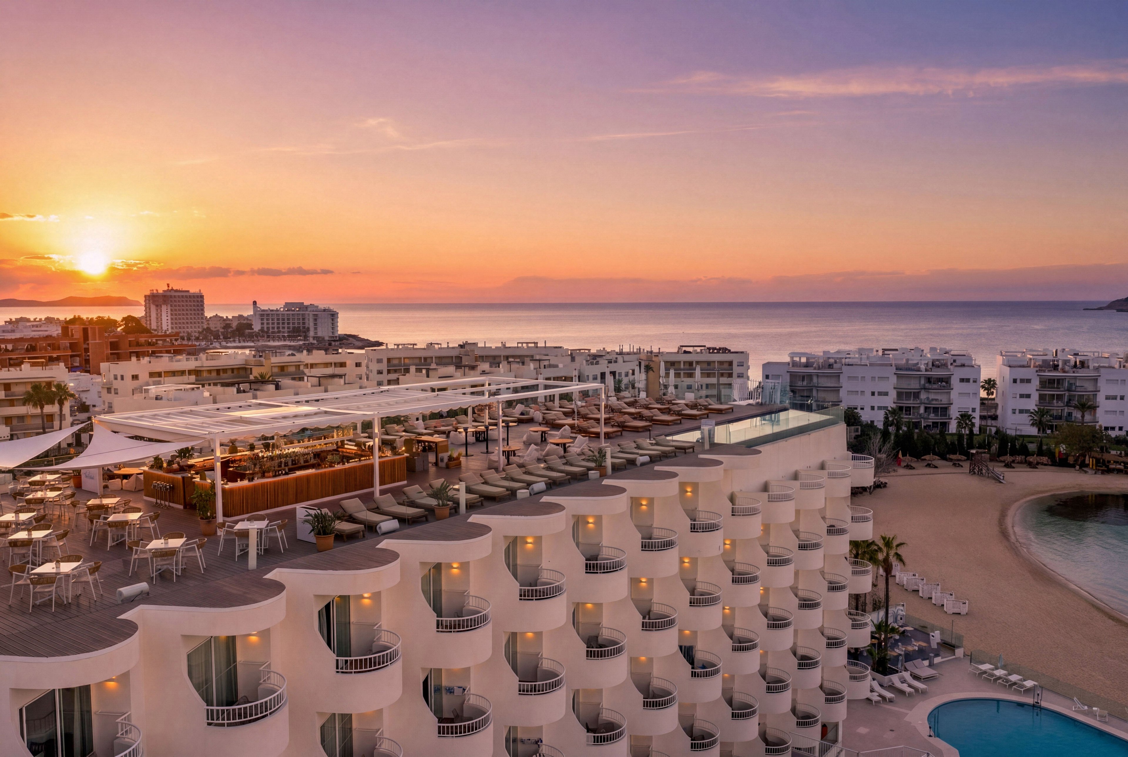 A modern seaside resort hotel overlooks a calm bay at sunset. The building features multiple floors of curved balconies facing the ocean and a large rooftop terrace with seating and shade structures. In the background, low-rise coastal buildings line the shoreline, while the sun sets over the distant horizon. The sky glows with warm orange and pink tones, reflecting softly on the water and creating a tranquil vacation atmosphere.