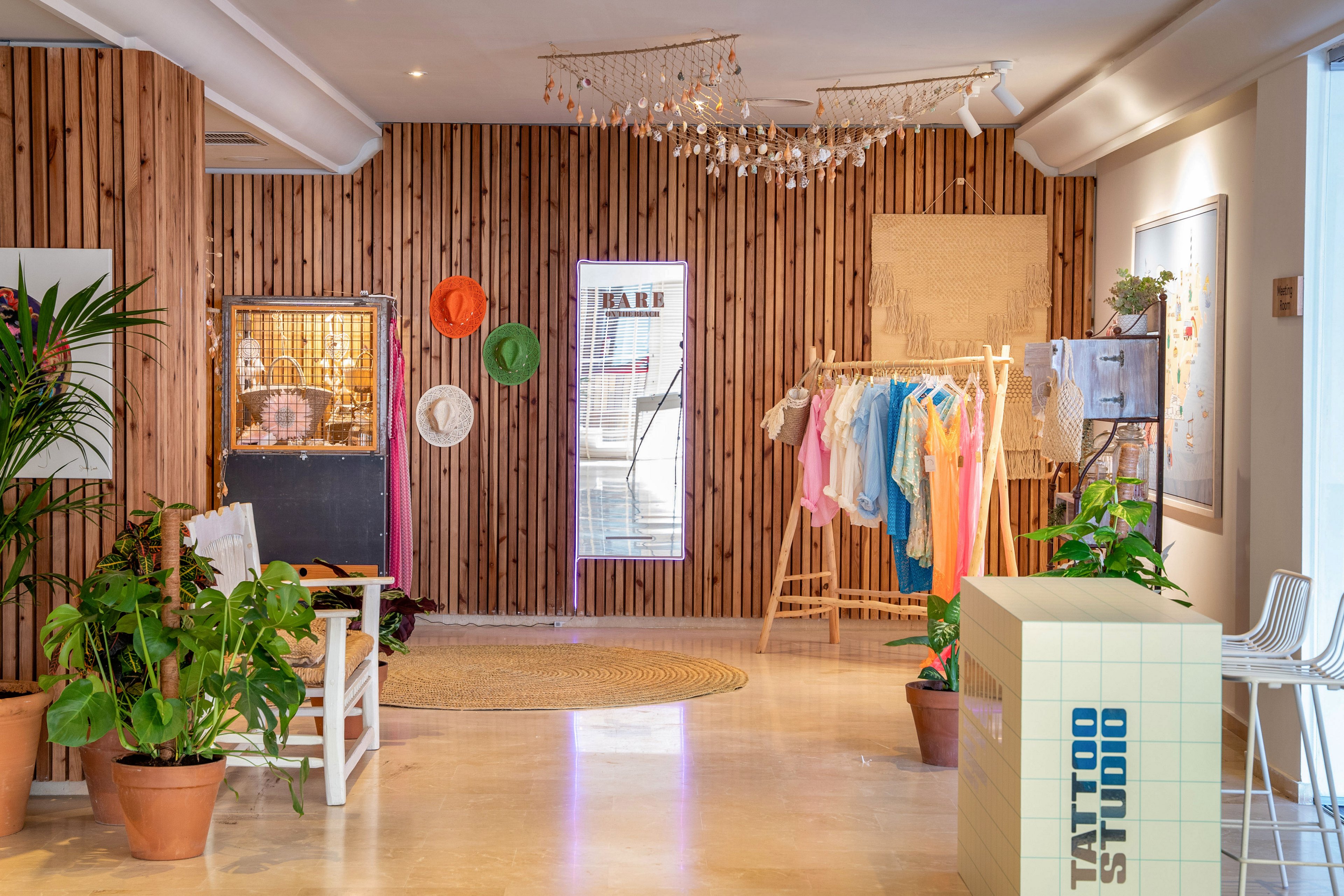 A stylish boutique interior features a wooden slat wall and a central display of colorful clothing on a rack. Sun hats and decorative items hang on the wall, while a glass display case and a door with a vertical window add depth to the space. Potted plants and a woven rug contribute to a relaxed, tropical atmosphere. Soft lighting and pastel tones create a warm, inviting mood suitable for a modern retail environment.