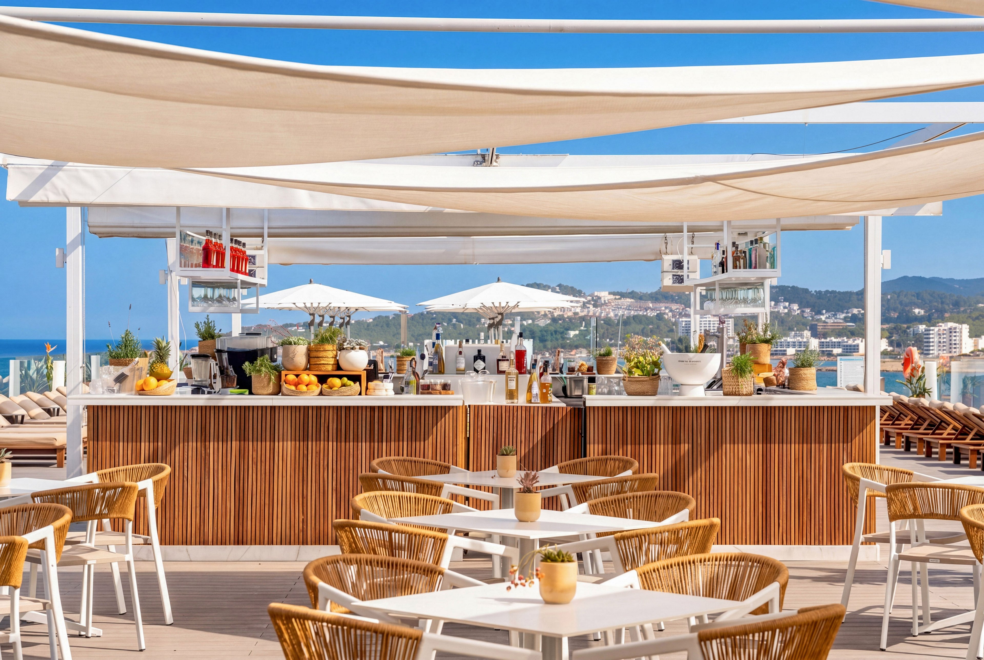 An open-air rooftop bar features a central counter stocked with fresh fruits, bottles, and glassware. White tables with wicker chairs are arranged neatly in the foreground under stretched fabric canopies. Large white umbrellas shade additional seating while the coastline and cityscape are visible in the distance. The bright daylight and neutral tones create a relaxed, summery atmosphere ideal for outdoor dining and drinks.