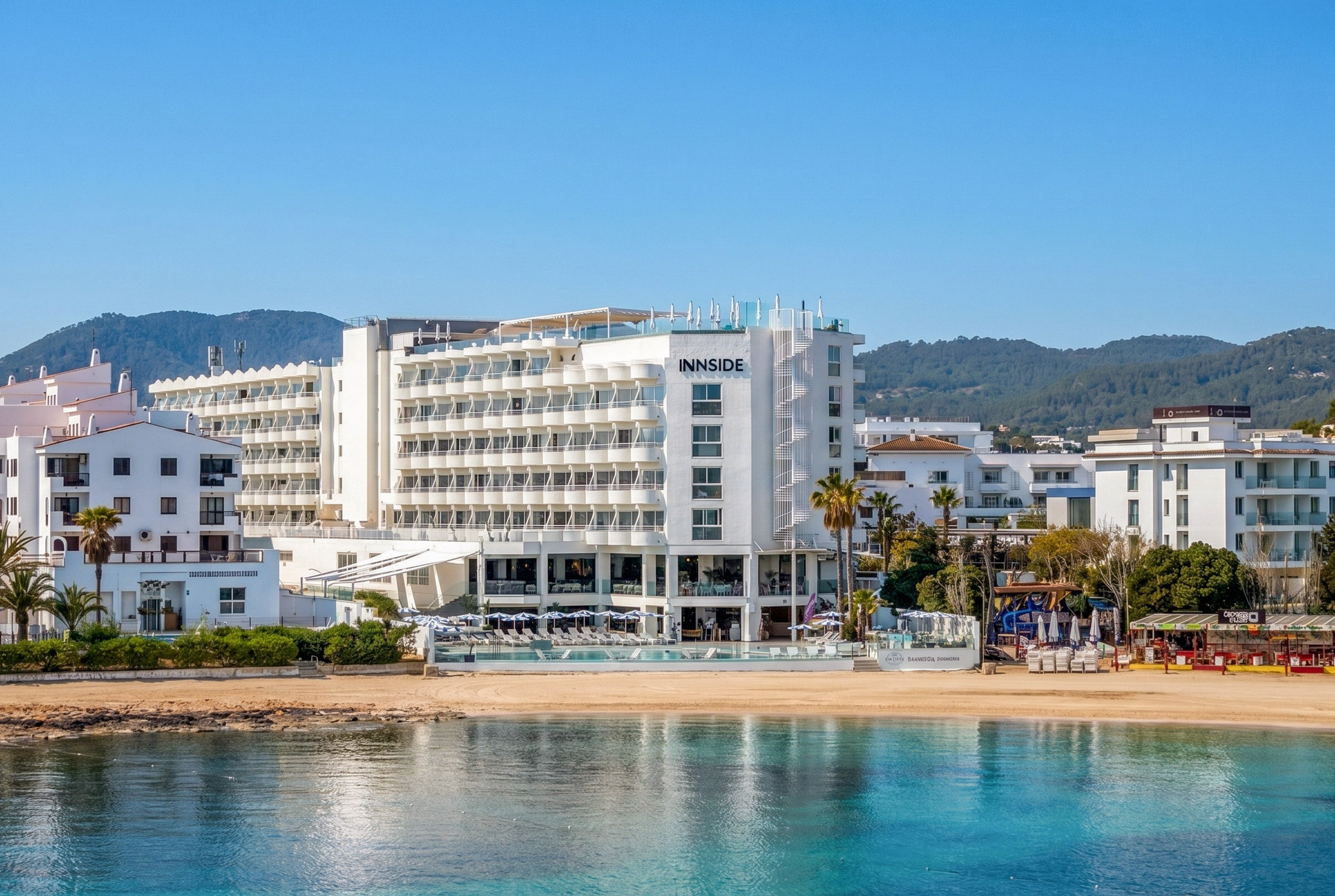 A contemporary multi-story hotel stands directly on a sandy beachfront beside clear blue water. The building features long balconies, glass railings, and a pool terrace facing the sea. Palm trees and nearby low-rise buildings frame the resort, with green hills rising in the background. The scene is bright and sunny, suggesting a relaxed coastal vacation atmosphere.
