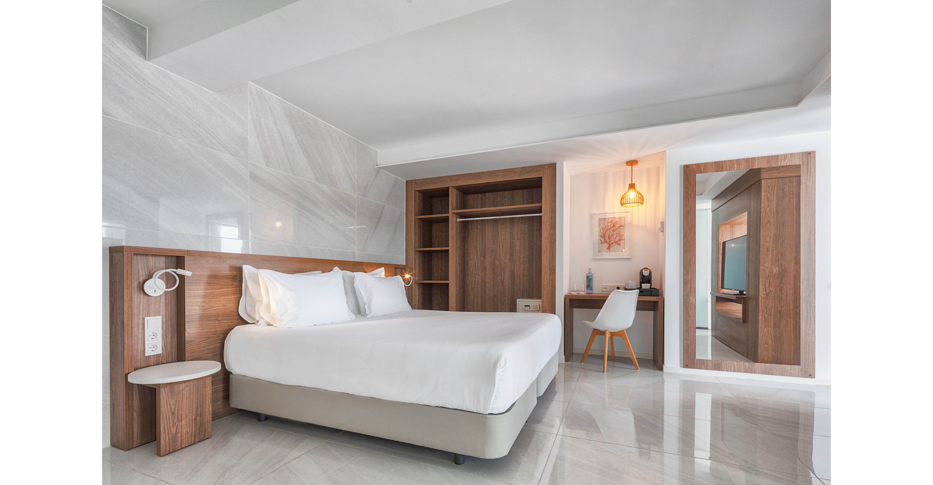 Contemporary hotel room features a neatly made double bed with white linens and a wooden headboard. The space includes an open wardrobe, a wall-mounted desk with a white chair, and a large framed mirror. Warm lighting from a hanging pendant lamp contrasts with the cool, glossy tiled floor and light gray walls. Overall, the room feels bright, clean, and inviting for travelers or business guests.