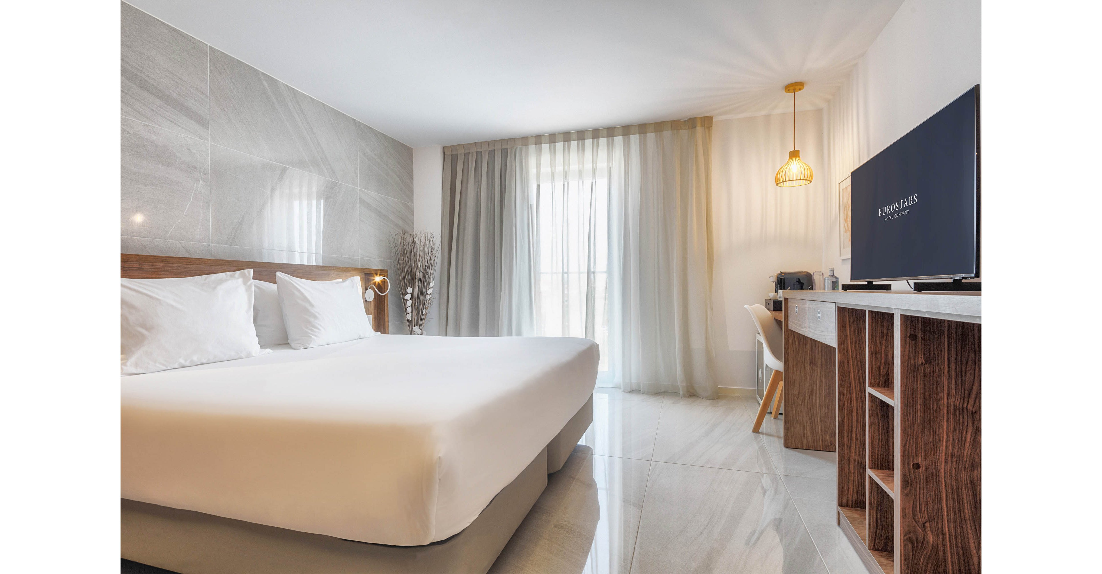 A contemporary hotel bedroom features a large neatly made bed with white linens and pillows. The room includes a wooden desk with a flat-screen television and a simple chair, creating a functional workspace. Floor-to-ceiling sheer curtains cover a large window, allowing soft natural light to fill the space. Neutral tones, glossy flooring, and minimal decor contribute to a clean, modern, and relaxing atmosphere.