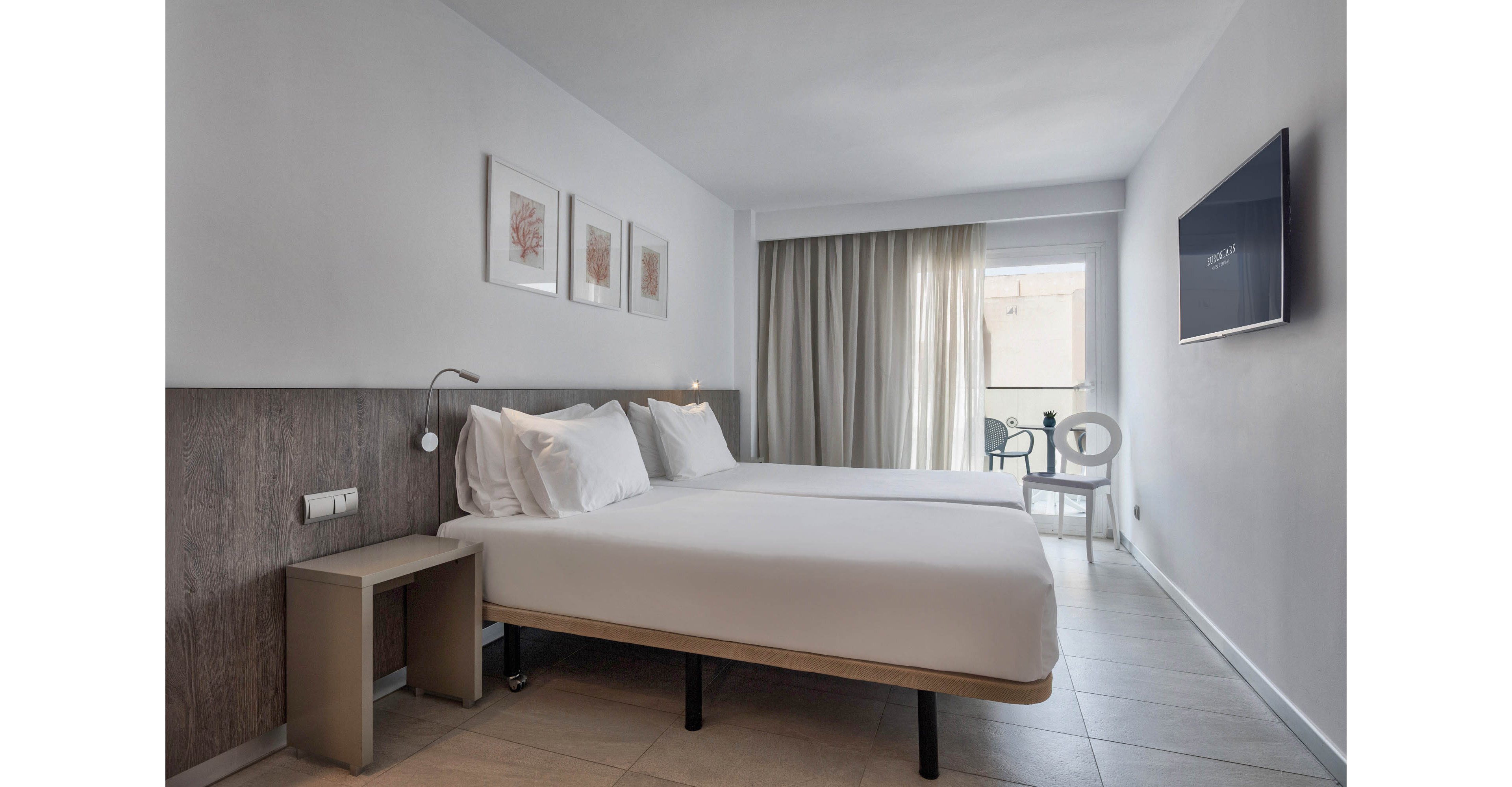 A contemporary hotel room features a large neatly made bed with crisp white linens. The space is decorated in a minimalist style with light walls, simple artwork, and a wooden headboard. A flat-screen television is mounted on the wall opposite the bed, and a small bedside table stands nearby. Sheer curtains cover a glass door leading to a balcony with outdoor seating, allowing natural light to brighten the room.