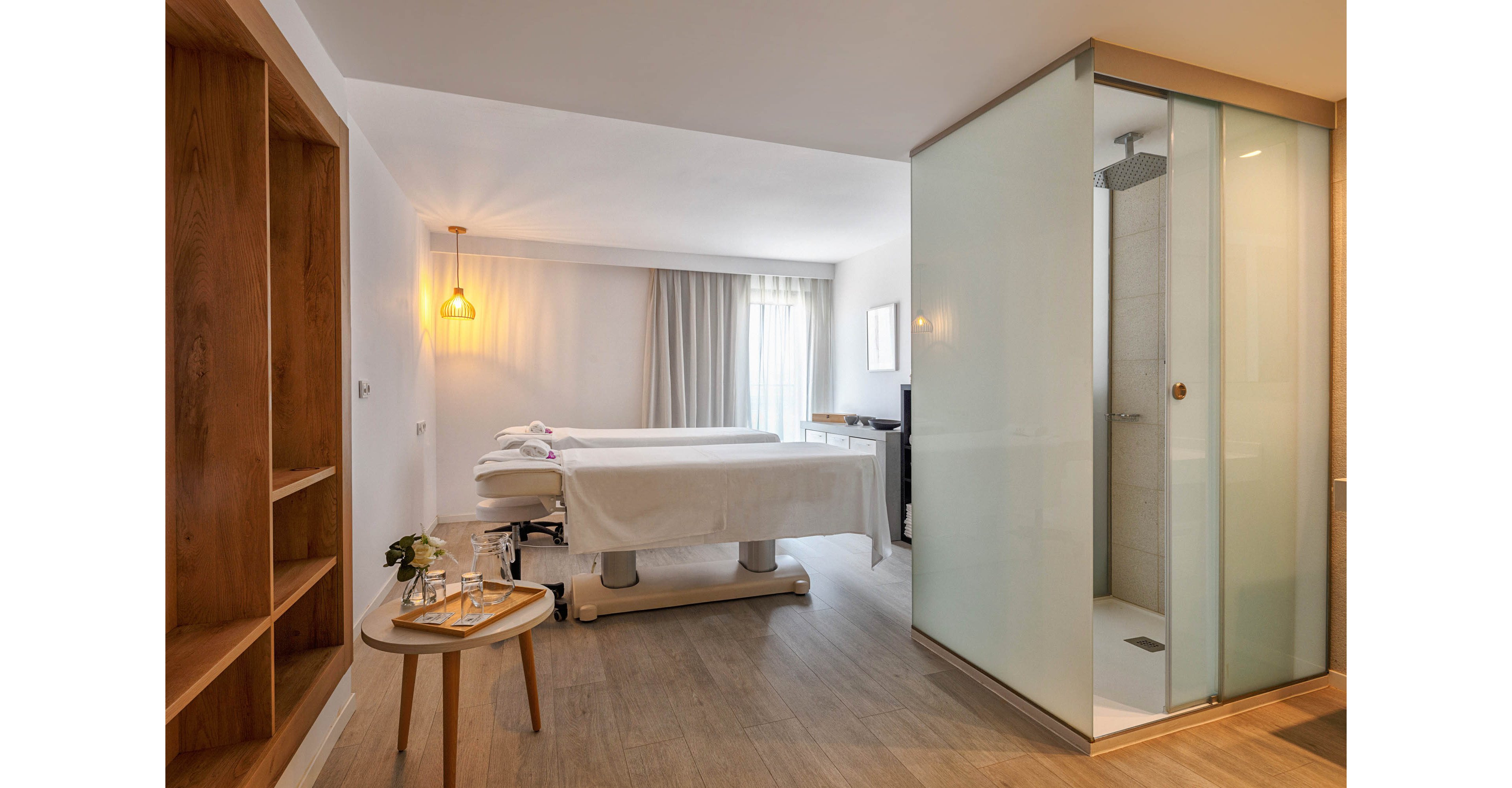 A contemporary spa treatment room features two massage tables neatly prepared with towels. The space is bright and airy, with large windows covered by light curtains allowing natural light to filter in. A glass-enclosed shower stands to the right, adding convenience and a modern touch. Wooden flooring, open shelving, and a small side table with water and flowers create a warm, relaxing atmosphere.