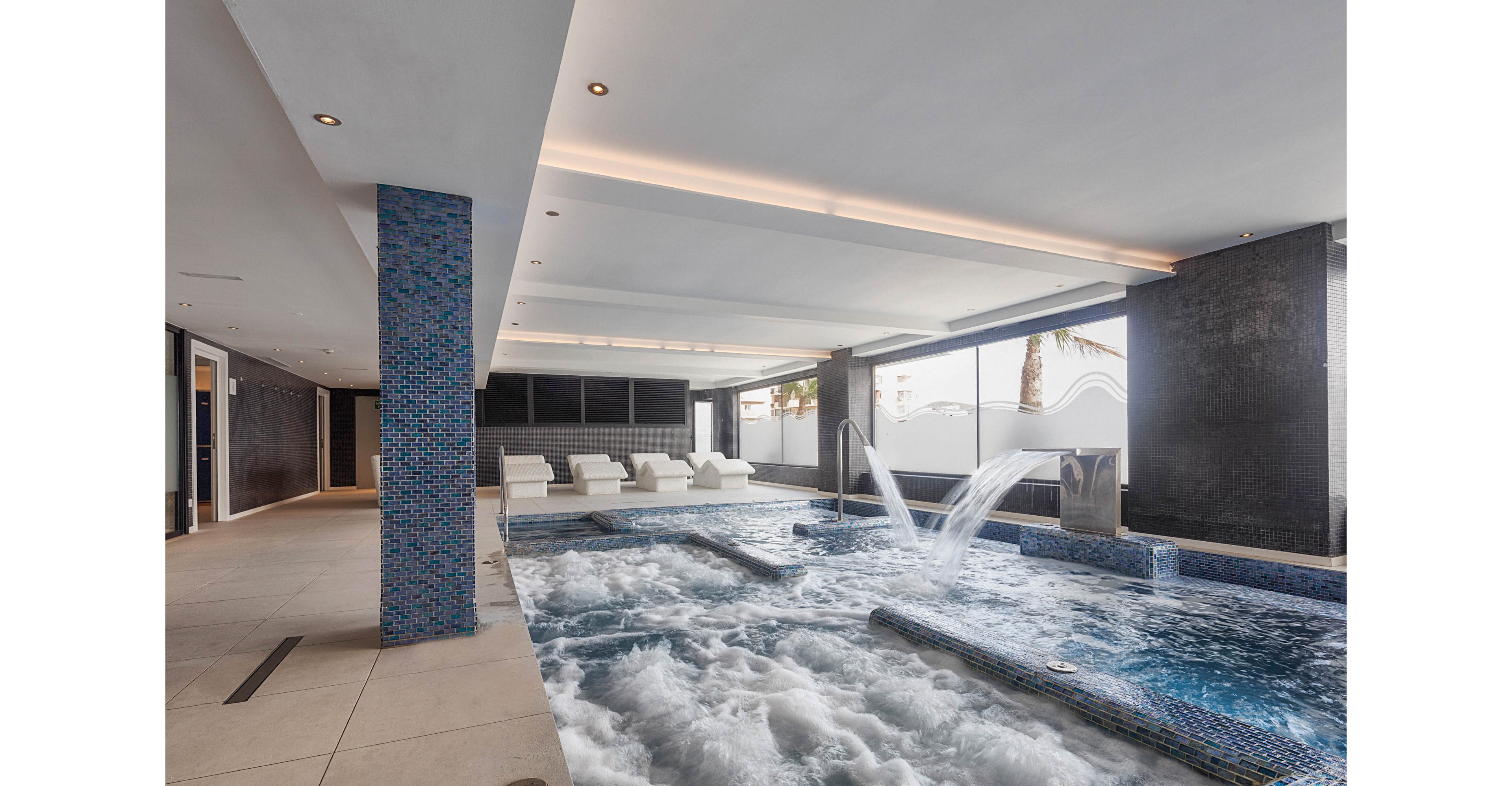 A contemporary indoor spa pool features powerful water jets and bubbling water. The space includes tiled columns and built-in loungers along one side of the pool. Large windows allow natural light to enter, revealing a glimpse of palm trees outside. The clean, minimalist design and neutral tones create a relaxing, luxurious atmosphere.