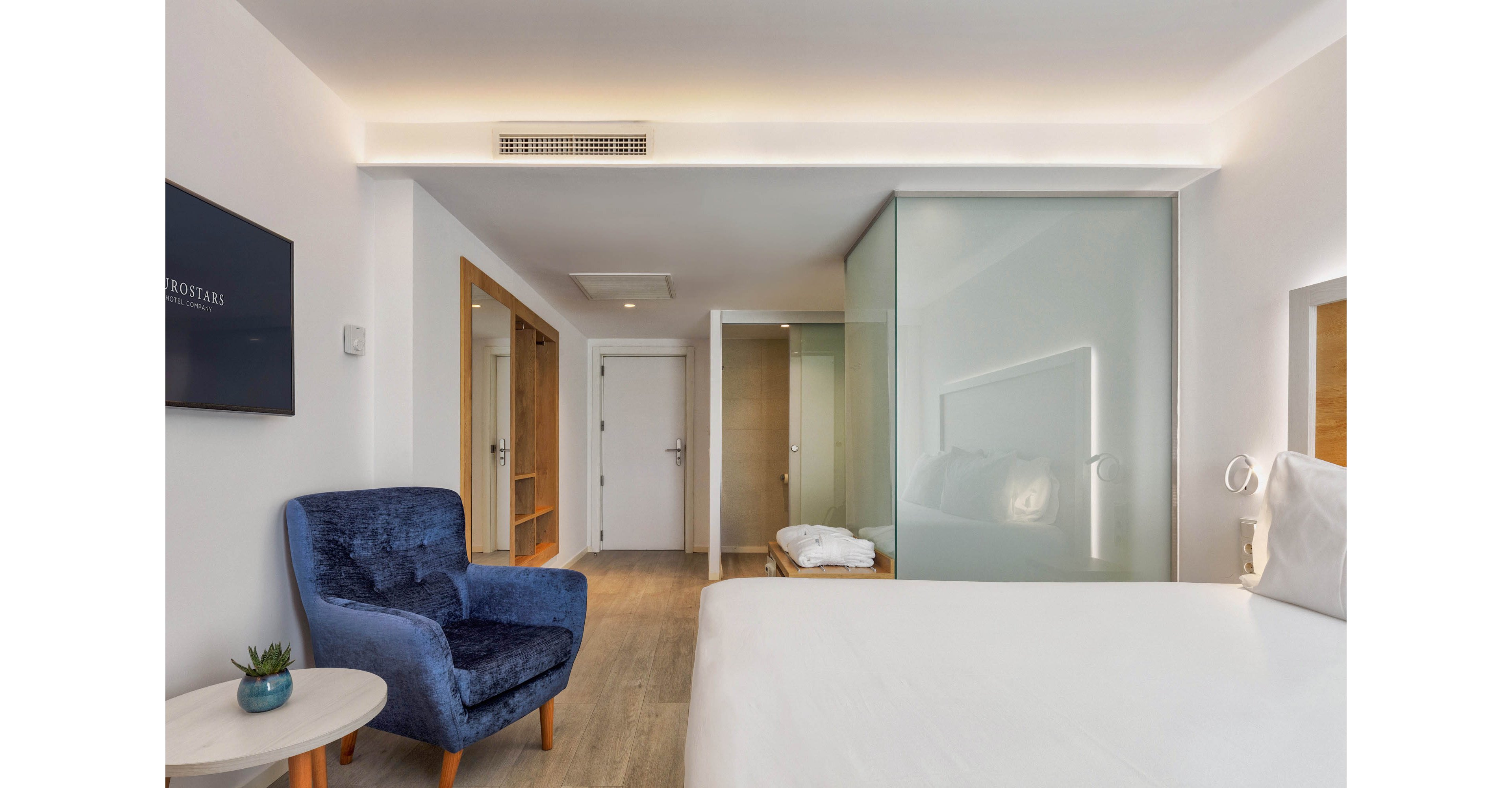Contemporary hotel room interior featuring a neatly made bed and a plush blue armchair beside a small round table. The space is bright with white walls, light wood flooring, and recessed ceiling lighting. A frosted glass partition separates the sleeping area from the bathroom, where folded white towels are visible. A wall-mounted television and built-in wardrobe complete the clean, minimalist design.
