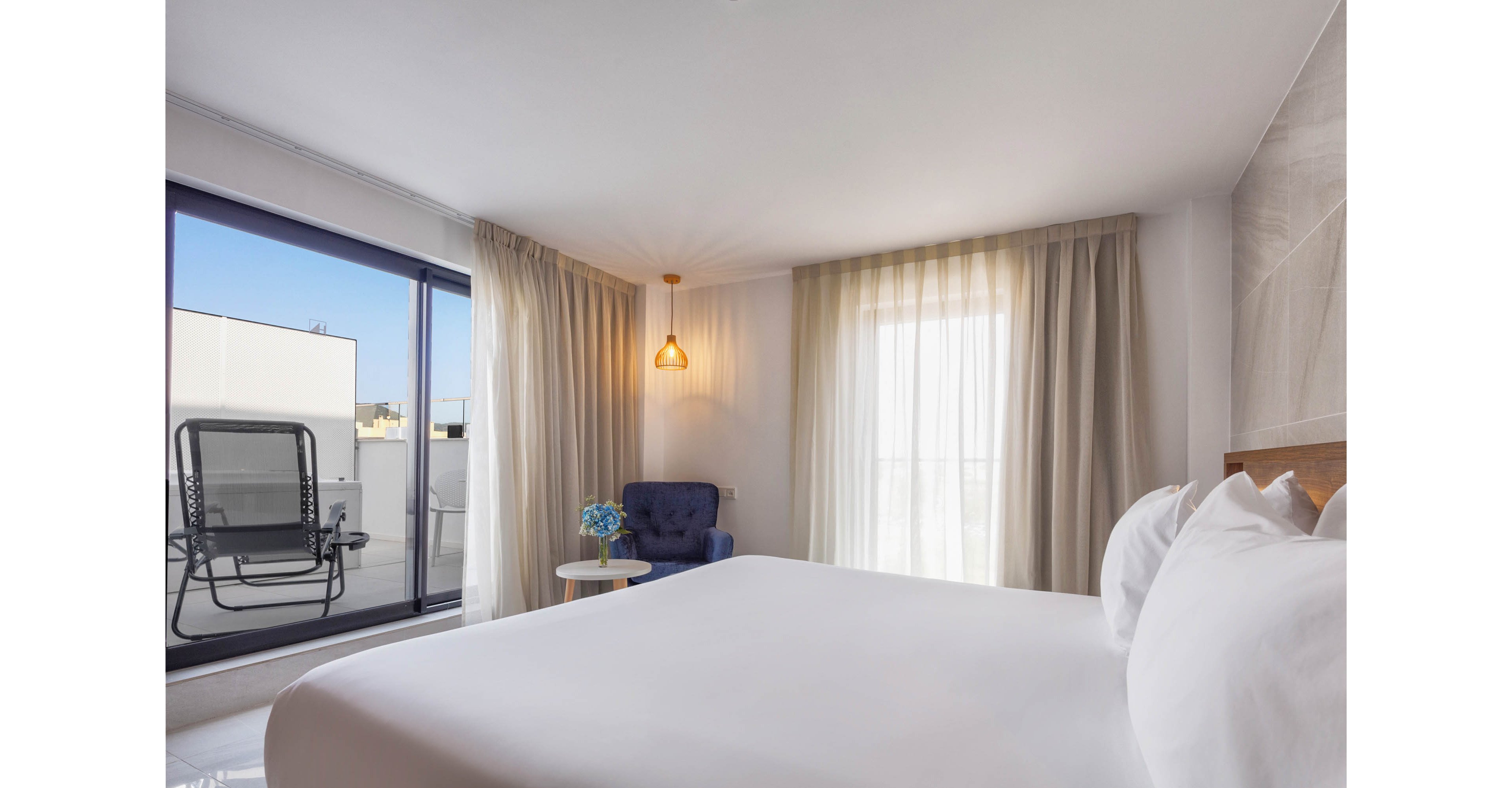 A bright modern hotel room features a large neatly made bed in the foreground. Floor-to-ceiling curtains filter natural light from a sliding glass door that opens onto a private terrace with outdoor seating. A blue armchair, small side table, and warm pendant light create a cozy corner inside the room. The overall atmosphere is clean, minimal, and inviting, with soft neutral tones and simple contemporary decor.