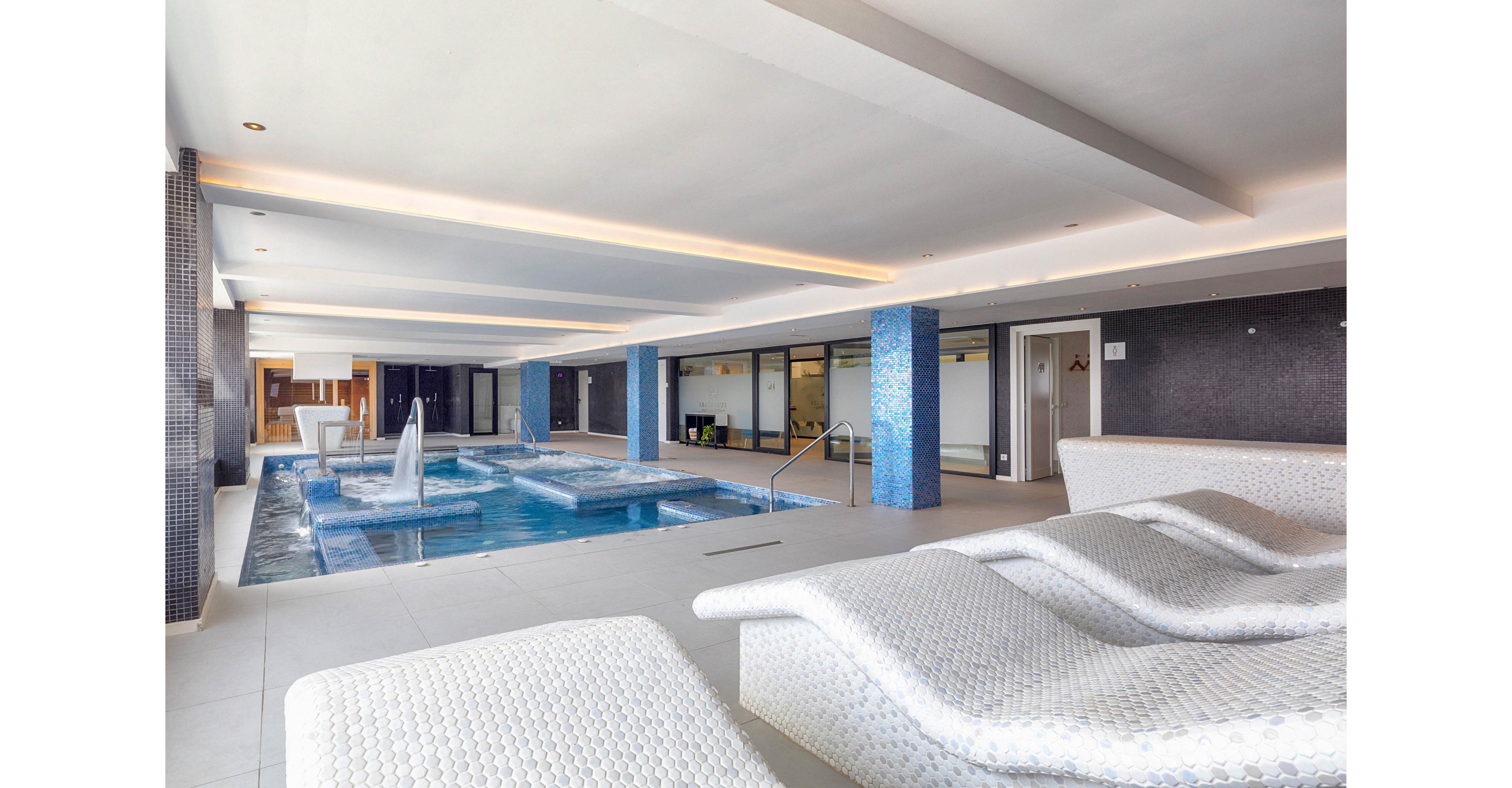 Contemporary indoor spa area featuring a rectangular pool with multiple water jets and fountains. White textured lounge chairs line the foreground, facing the pool for relaxation. Blue mosaic tiles accent the pool columns and edges, contrasting with the neutral walls and ceiling. Large windows and glass partitions in the background suggest a connected wellness or hotel facility.