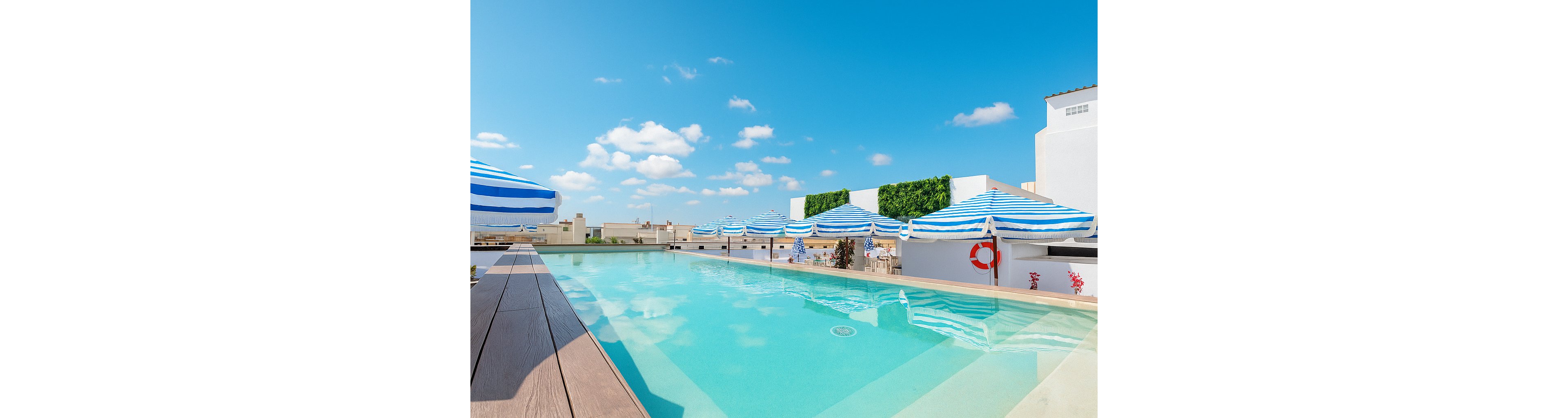 A clear rooftop swimming pool stretches alongside a wooden deck under a bright blue sky. Blue and white striped umbrellas line the far side of the pool, providing shade over lounge chairs. White buildings and a few green hedges frame the scene, suggesting an urban or resort setting. The calm water and scattered clouds create a relaxed, vacation-like atmosphere.