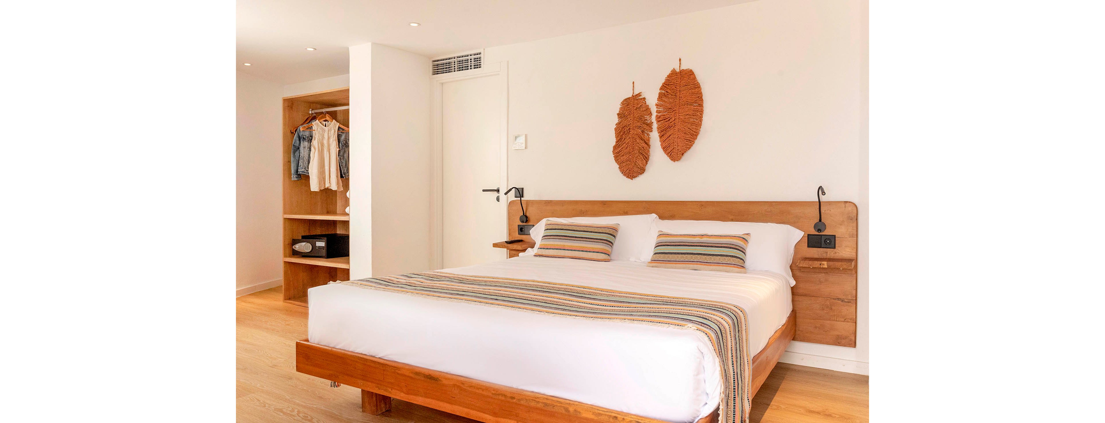 A bright, minimalist bedroom features a large wooden bed with crisp white linens and striped accent pillows. The room includes an open wooden wardrobe with neatly hung clothes and folded items. Two decorative leaf-shaped wall hangings are placed above the headboard, and modern bedside lights flank the bed. Light wood flooring and white walls create an airy, contemporary atmosphere.