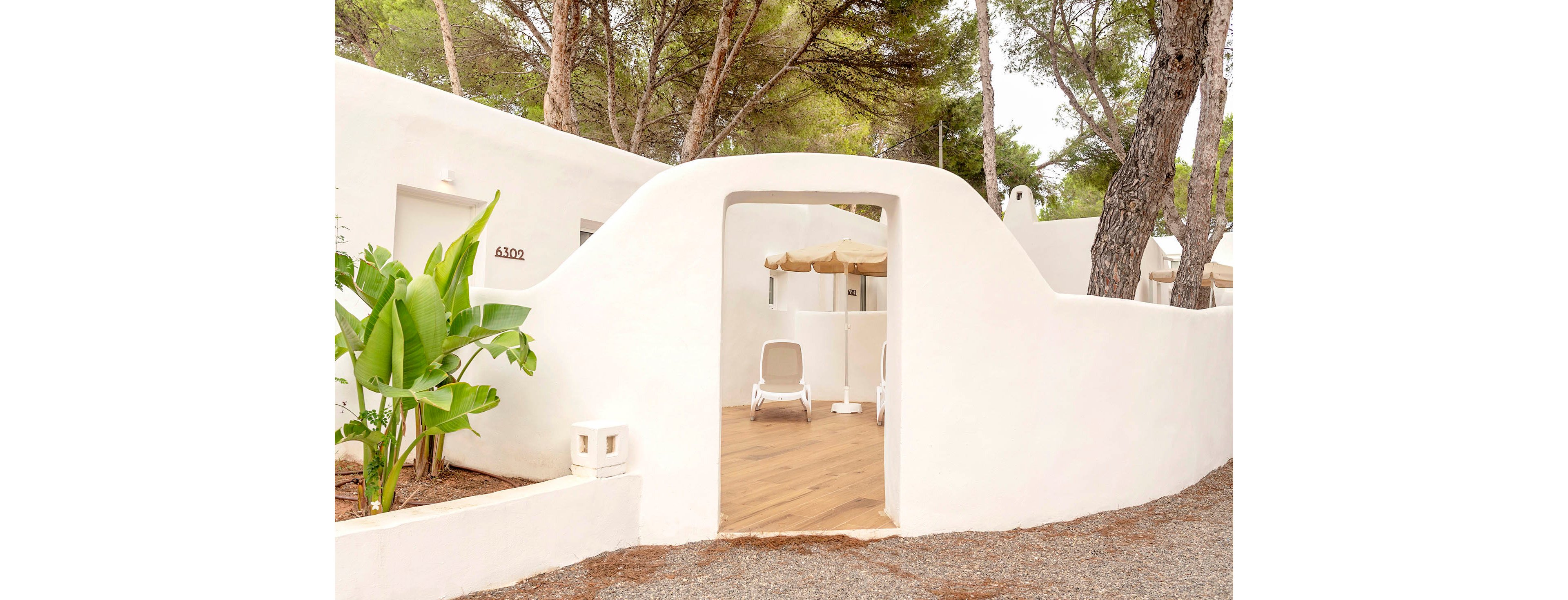 A minimalist white villa features a small enclosed patio framed by smooth, curved walls. Inside the courtyard, two white lounge chairs sit on a wooden deck beneath a beige outdoor umbrella. Lush green plants and tall pine trees surround the building, creating a tranquil, Mediterranean-style setting. The soft neutral tones and clean architectural lines convey a calm, relaxing holiday atmosphere.