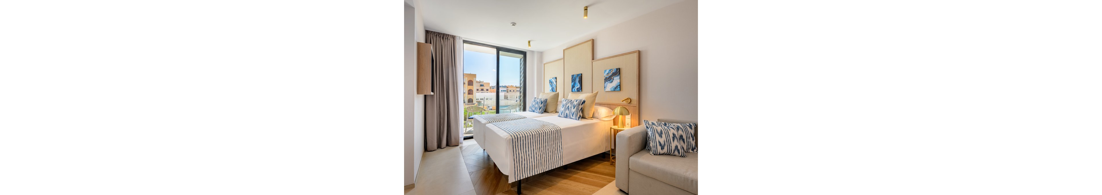 A contemporary hotel room features two neatly made twin beds with striped runners and patterned pillows. Large floor-to-ceiling windows open to a balcony overlooking a sunny urban courtyard. Soft neutral tones are accented by blue abstract artwork above the beds and matching textiles. A light-colored sofa, bedside table, and warm ceiling lights complete the cozy, inviting interior.