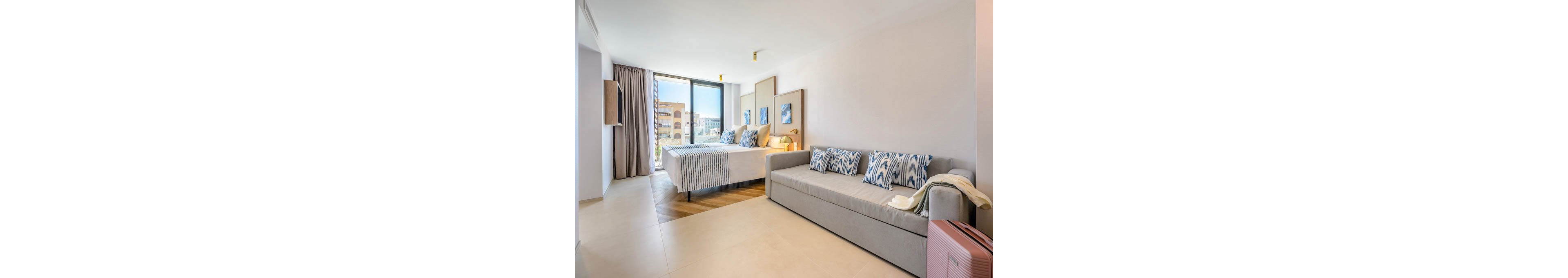 A contemporary hotel room features two twin beds with striped blue and white bedding and matching decorative pillows. A light gray sofa with patterned cushions sits opposite the beds, adding extra seating or sleeping space. Large floor-to-ceiling windows with curtains let in natural light and reveal nearby buildings outside. The room is decorated in a minimalist style with neutral walls, tiled flooring, and subtle coastal-inspired artwork above the beds.
