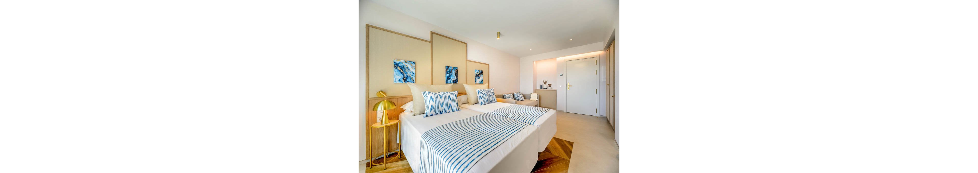 Contemporary twin beds with striped blue and white linens are arranged in a bright, minimalist hotel room. Abstract blue artwork hangs above the headboards, complementing patterned throw pillows. A small bedside table with a gold lamp and decorative objects adds warmth to the neutral walls and flooring. The space feels clean, airy, and inviting, with soft lighting and simple modern decor.